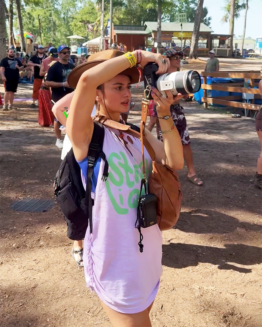 A person wearing a purple shirt uses a camera while shielding their eyes from the sun at an outdoor event.