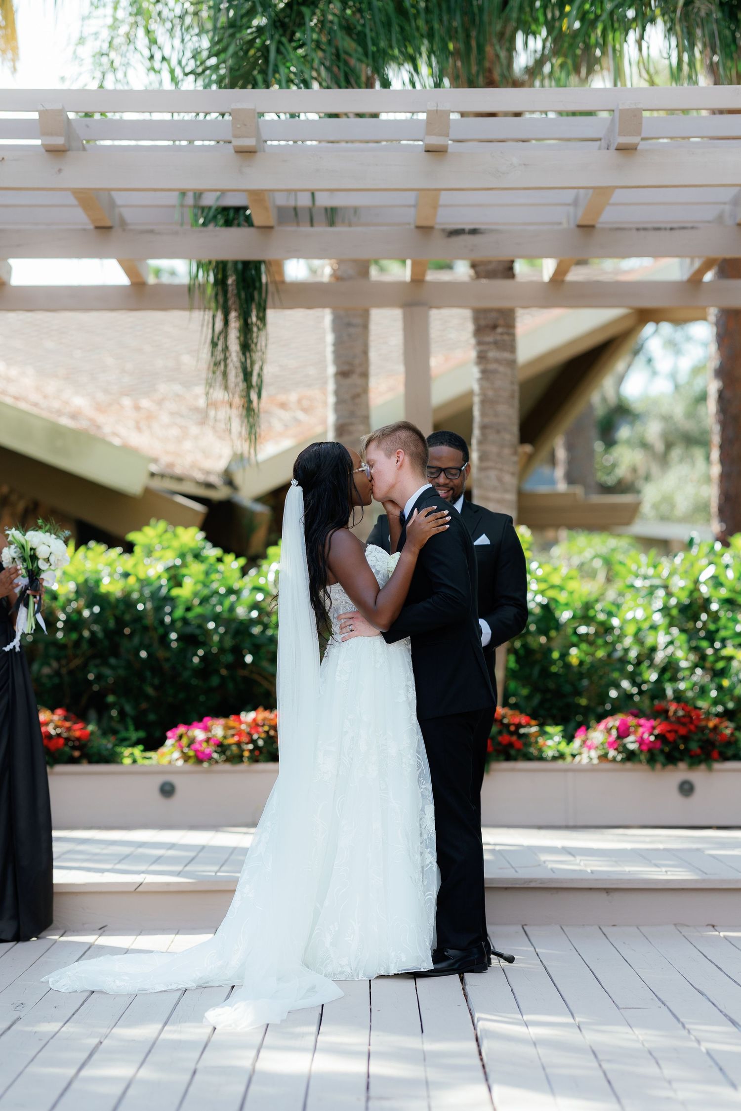 Interracial newlyweds share first kiss under wooden pergola surrounded by colorful flowers at outdoor ceremony.