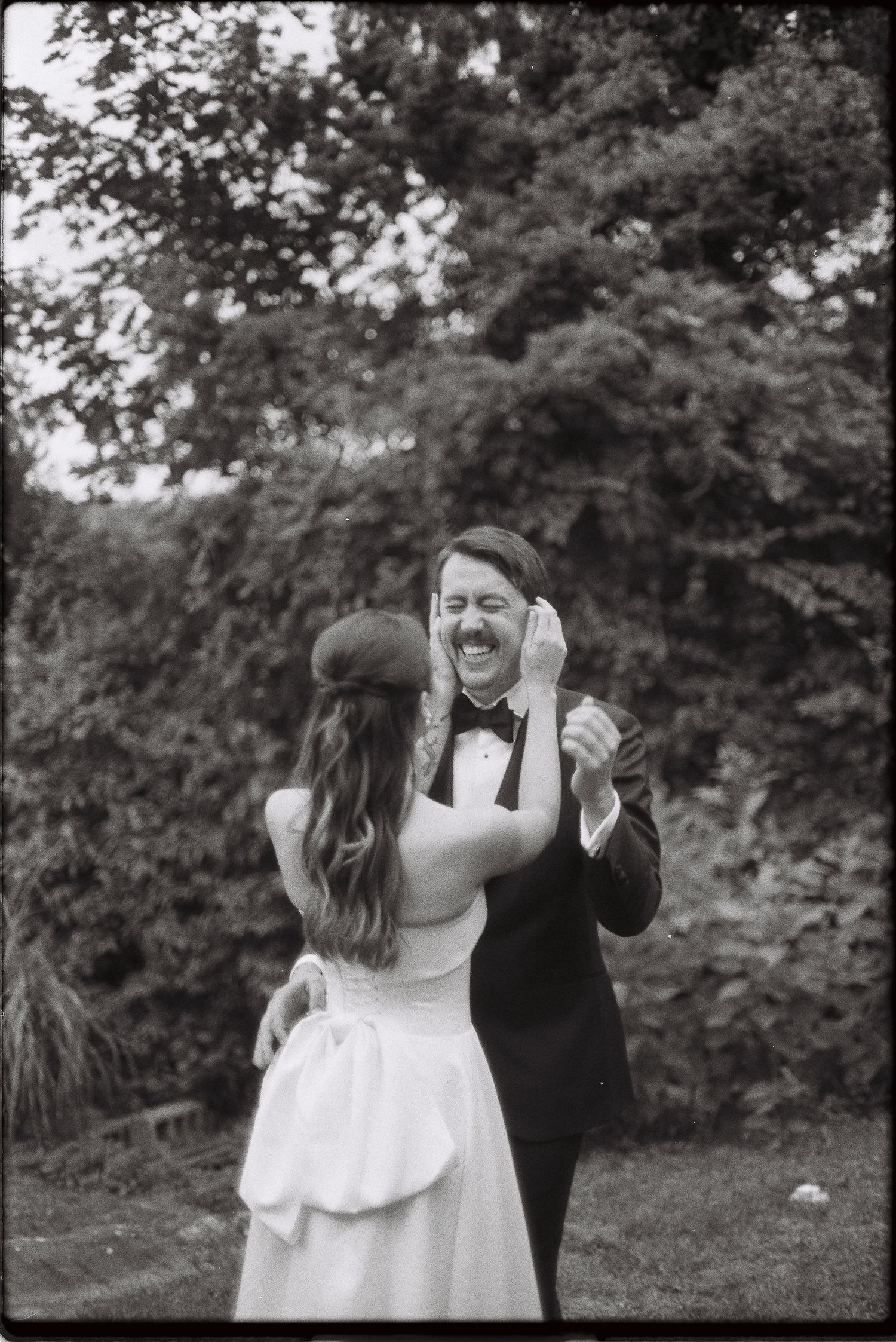 Black and white photo of joyful groom laughing as bride places hat on his head during outdoor reception.