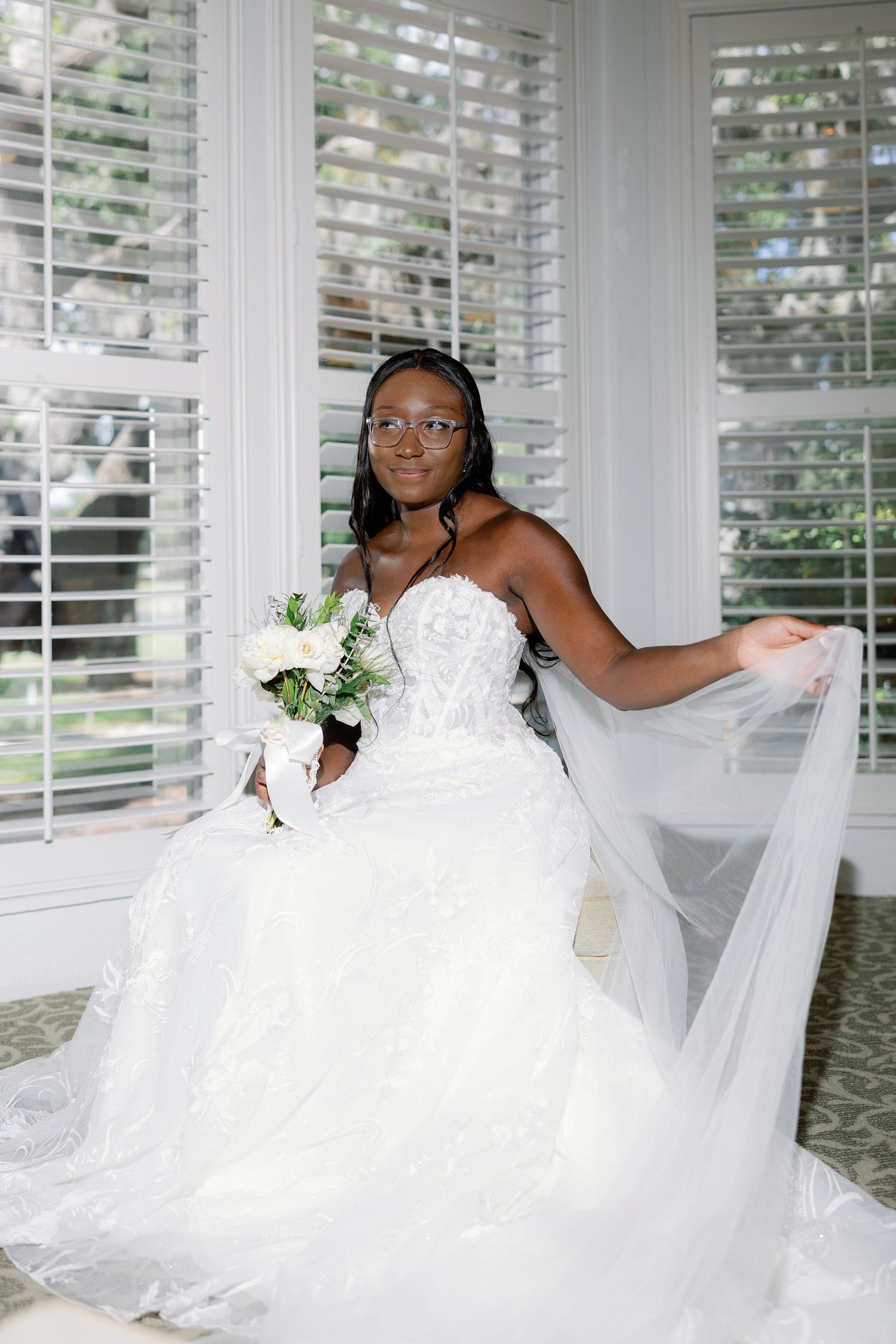 African American bride in beaded ball gown holds white bouquet while seated near window with veil.