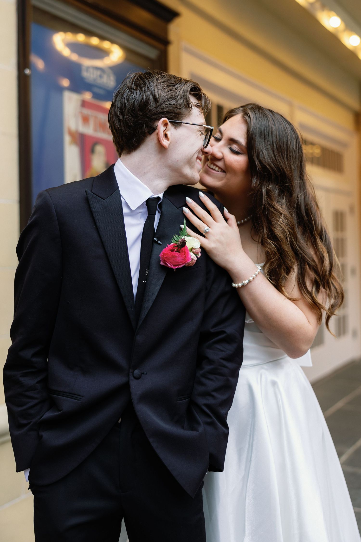 A groom in a black suit kisses his smiling bride in a white dress, who wears a bracelet and holds his lapel.