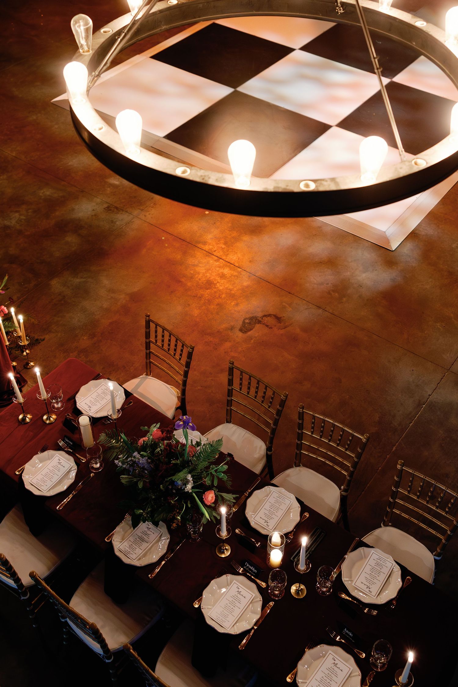 Aerial view of an elegant dining table set for a event beneath a circular chandelier with Edison bulbs.
