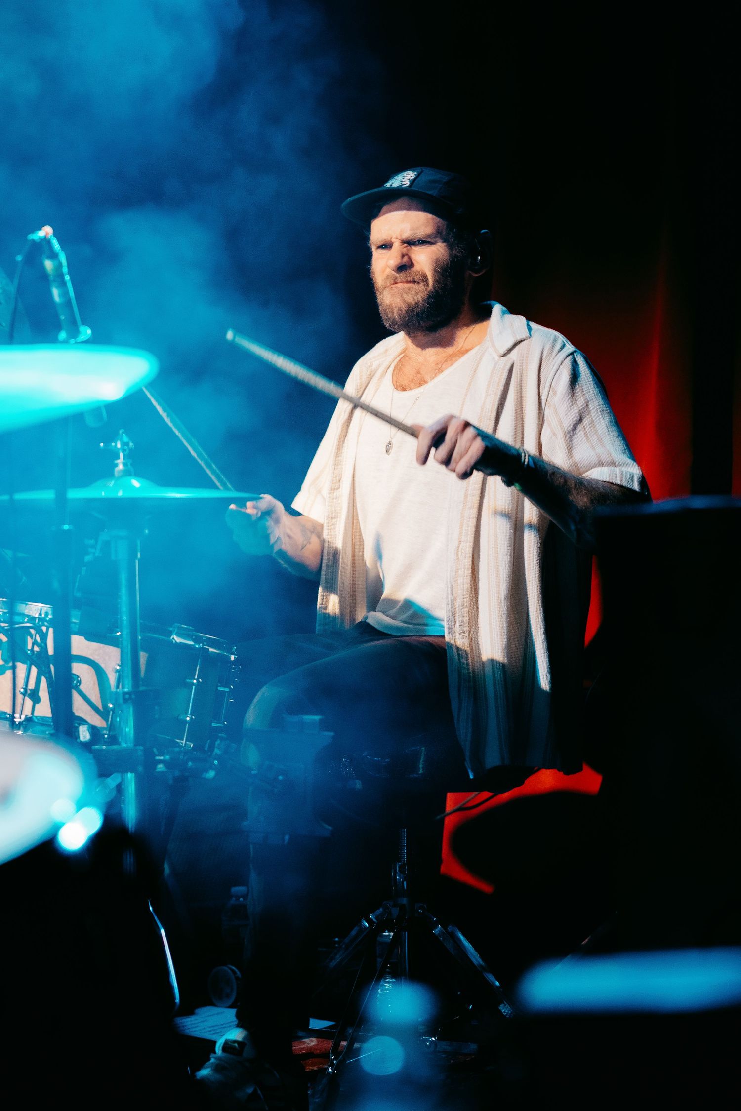 Drummer performs energetically on stage with dramatic blue lighting and smoke effects during a live concert performance.