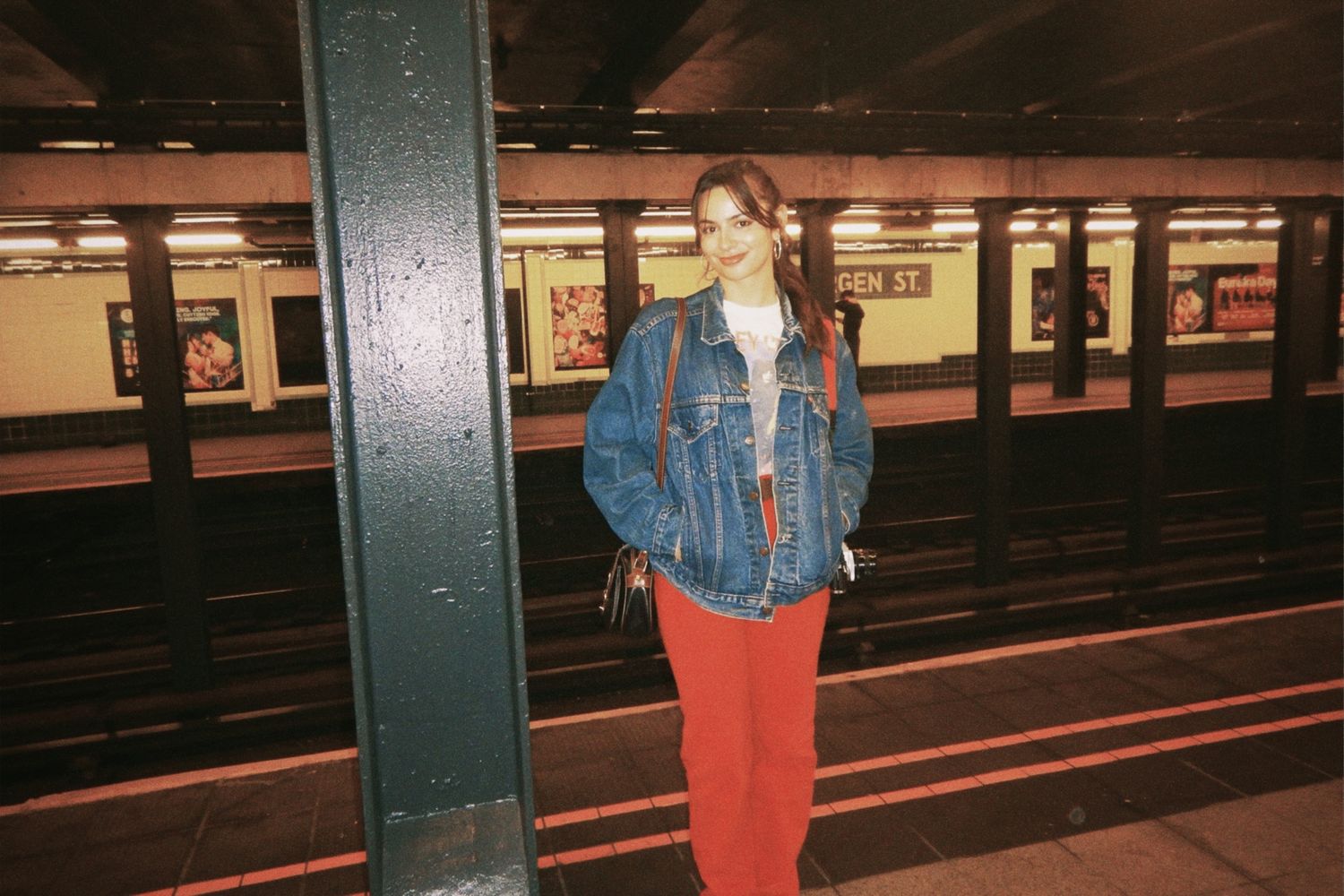 Person waiting alone on an empty subway platform near green support columns and dark train tracks.