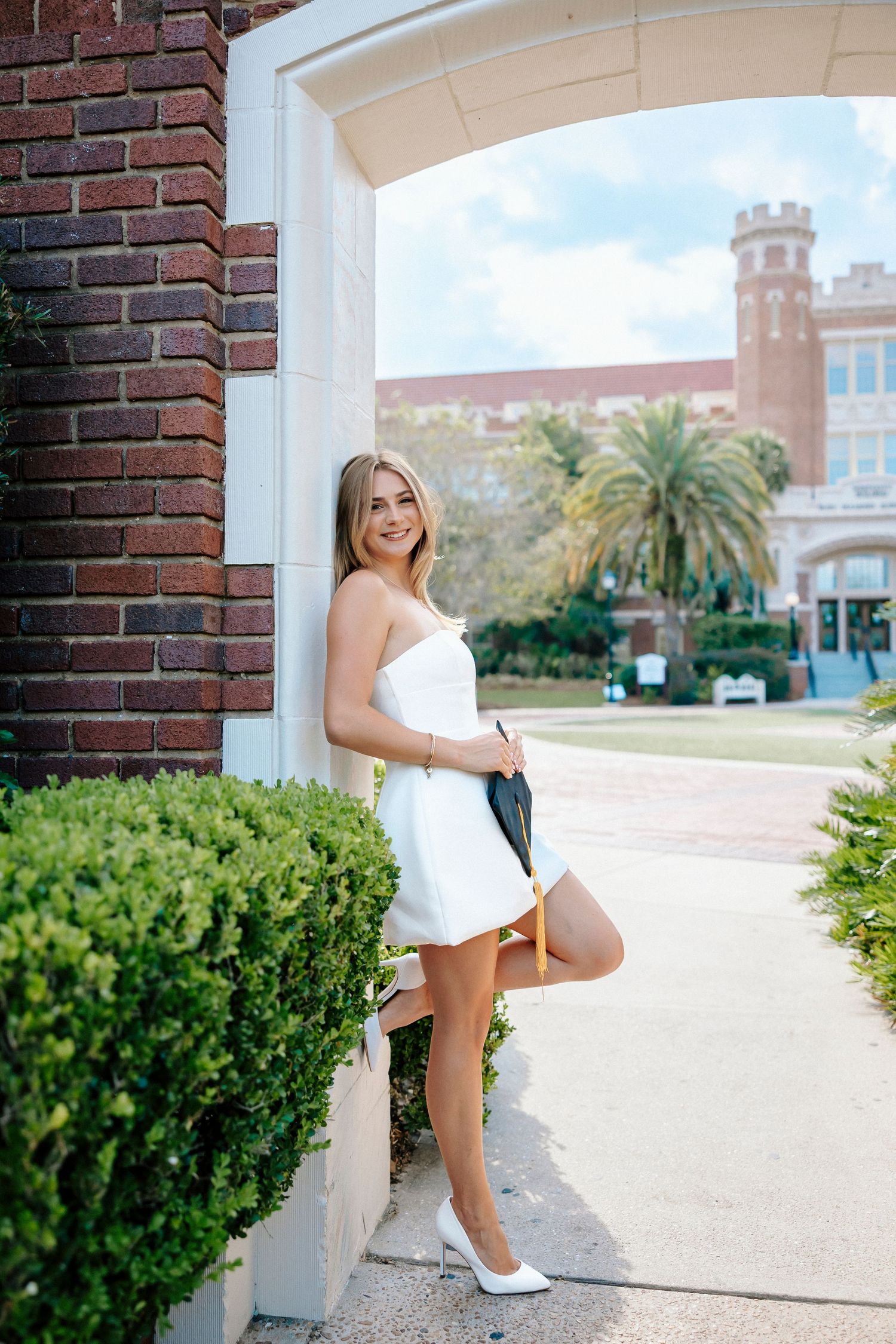 A figure in a white dress poses along a brick wall with palm trees and Spanish-style architecture in the background.