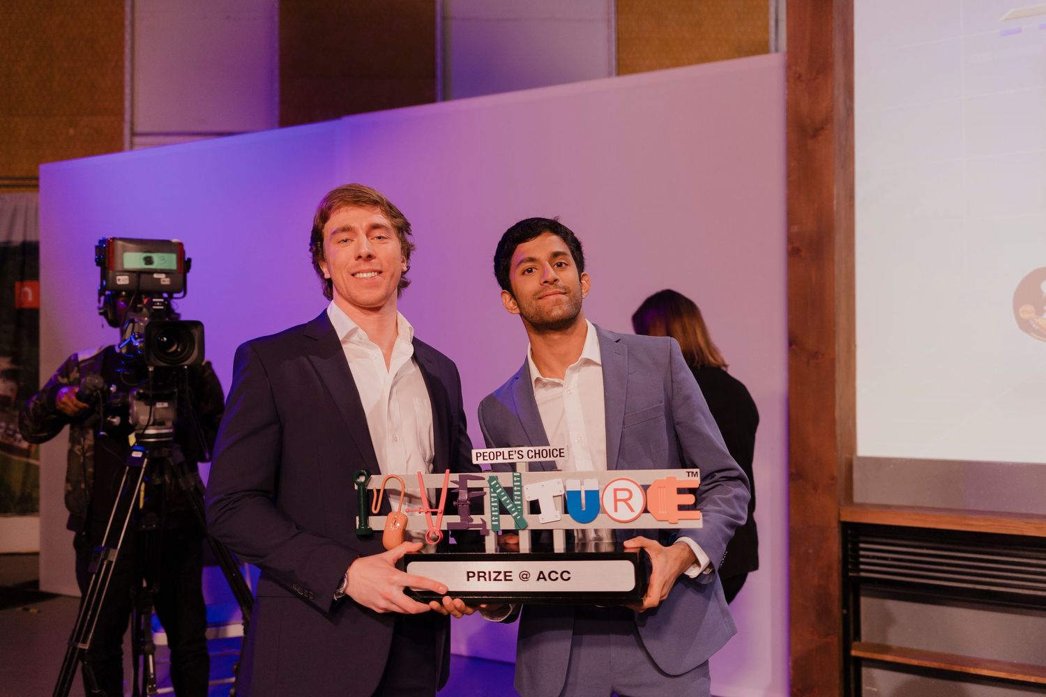 Two people in suits hold an award trophy at a presentation event with a video camera visible in the background.