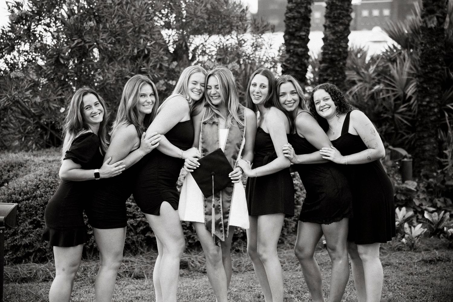 A group of women in black and white dresses pose together in an outdoor garden setting for a celebratory photo shoot.