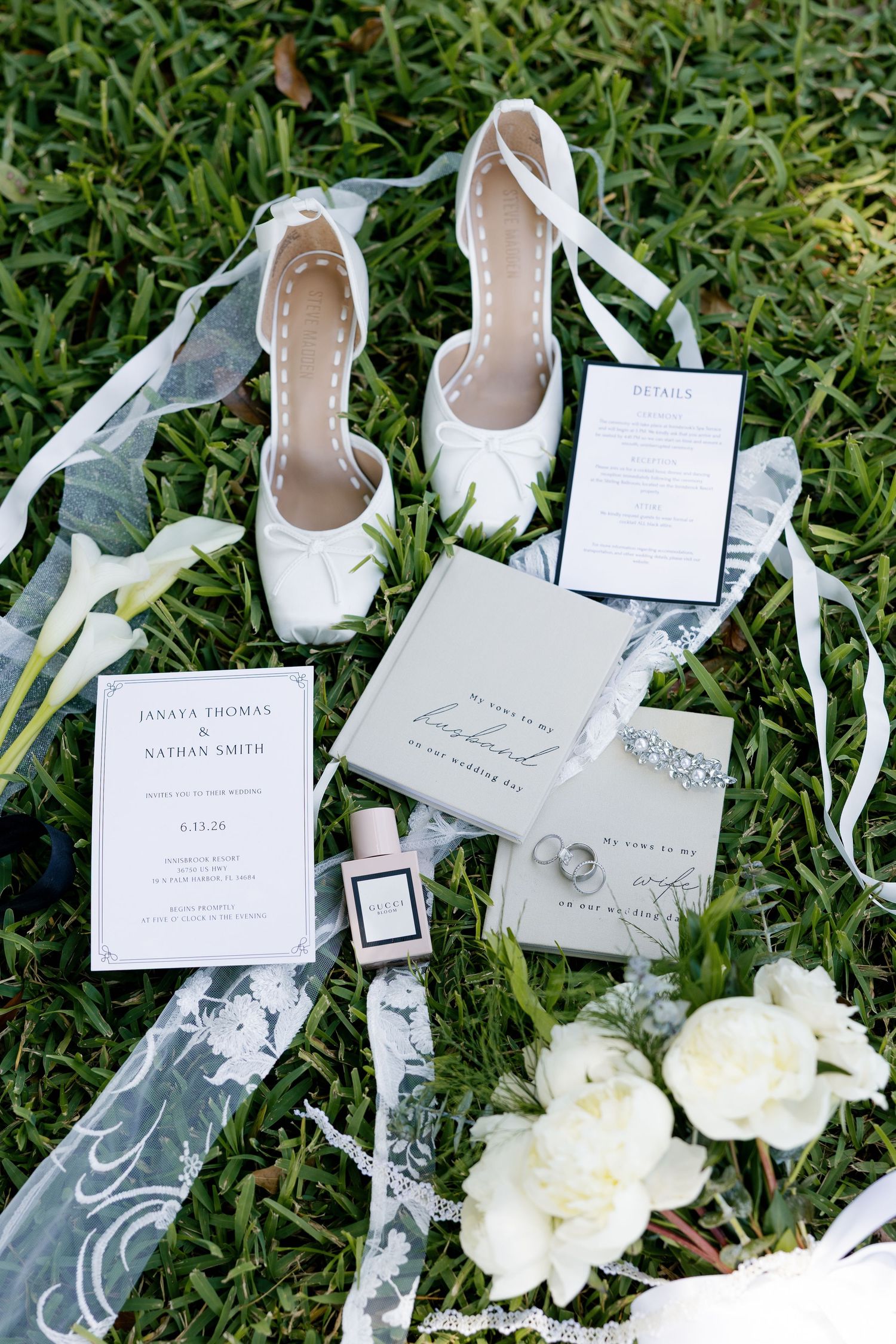 Elegant wedding flat lay featuring white bridal shoes, invitations, perfume, rings, and white peony bouquet on green grass.