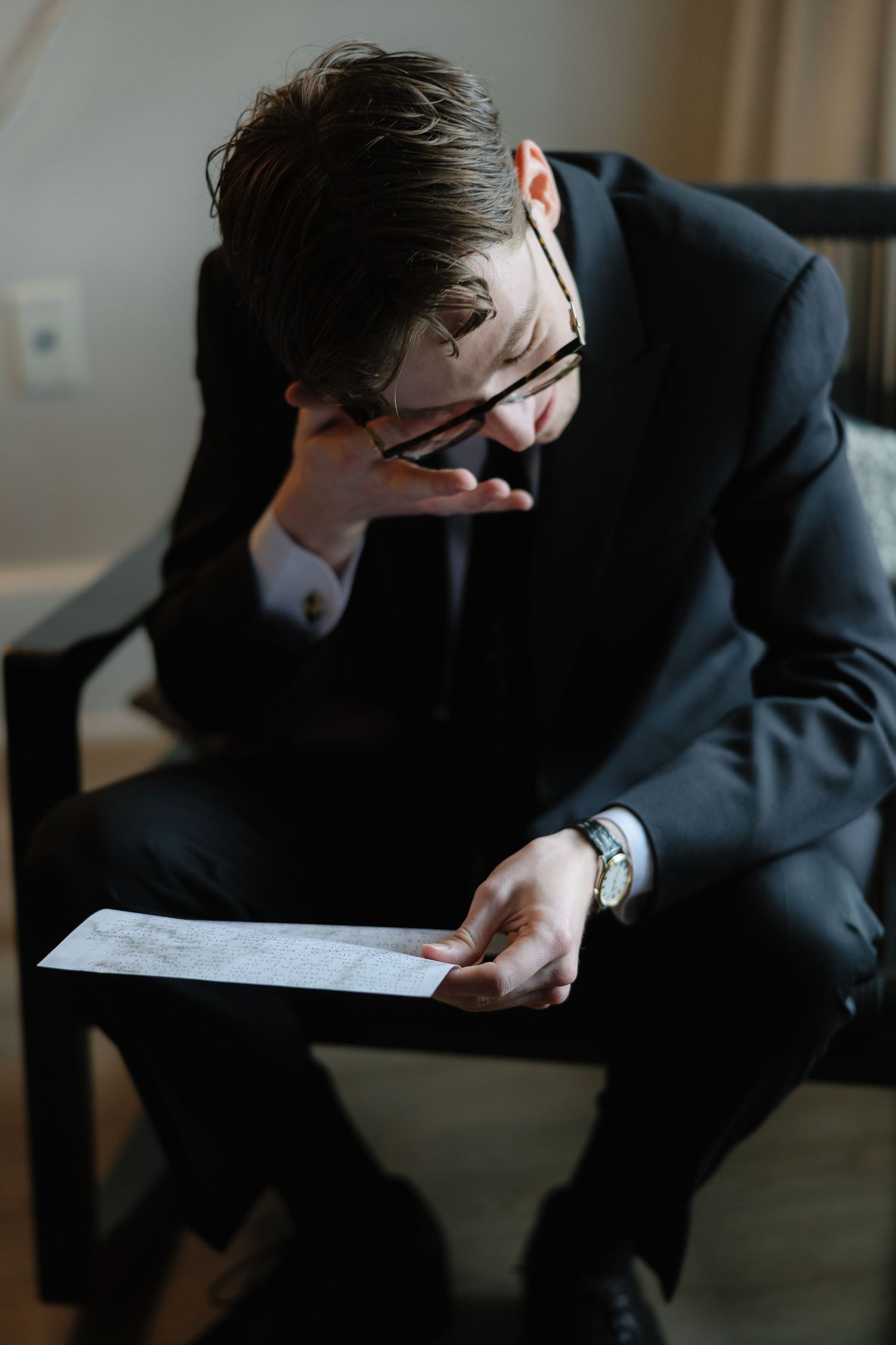 Stressed businessman in black suit reading document, adjusting glasses while seated in dark wooden chair.