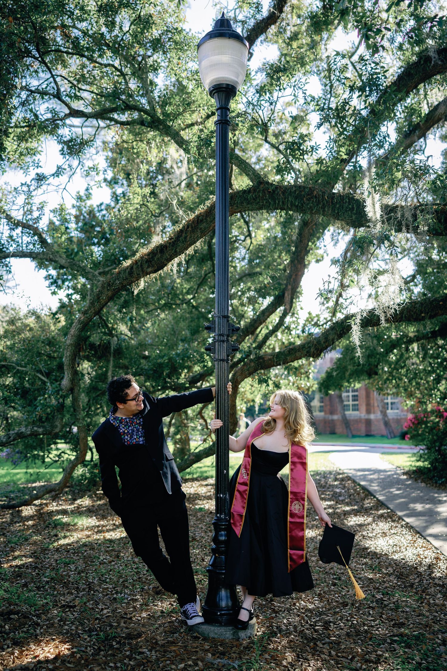 A couple in graduation regalia share playful moments under moss-draped trees on a college campus path.