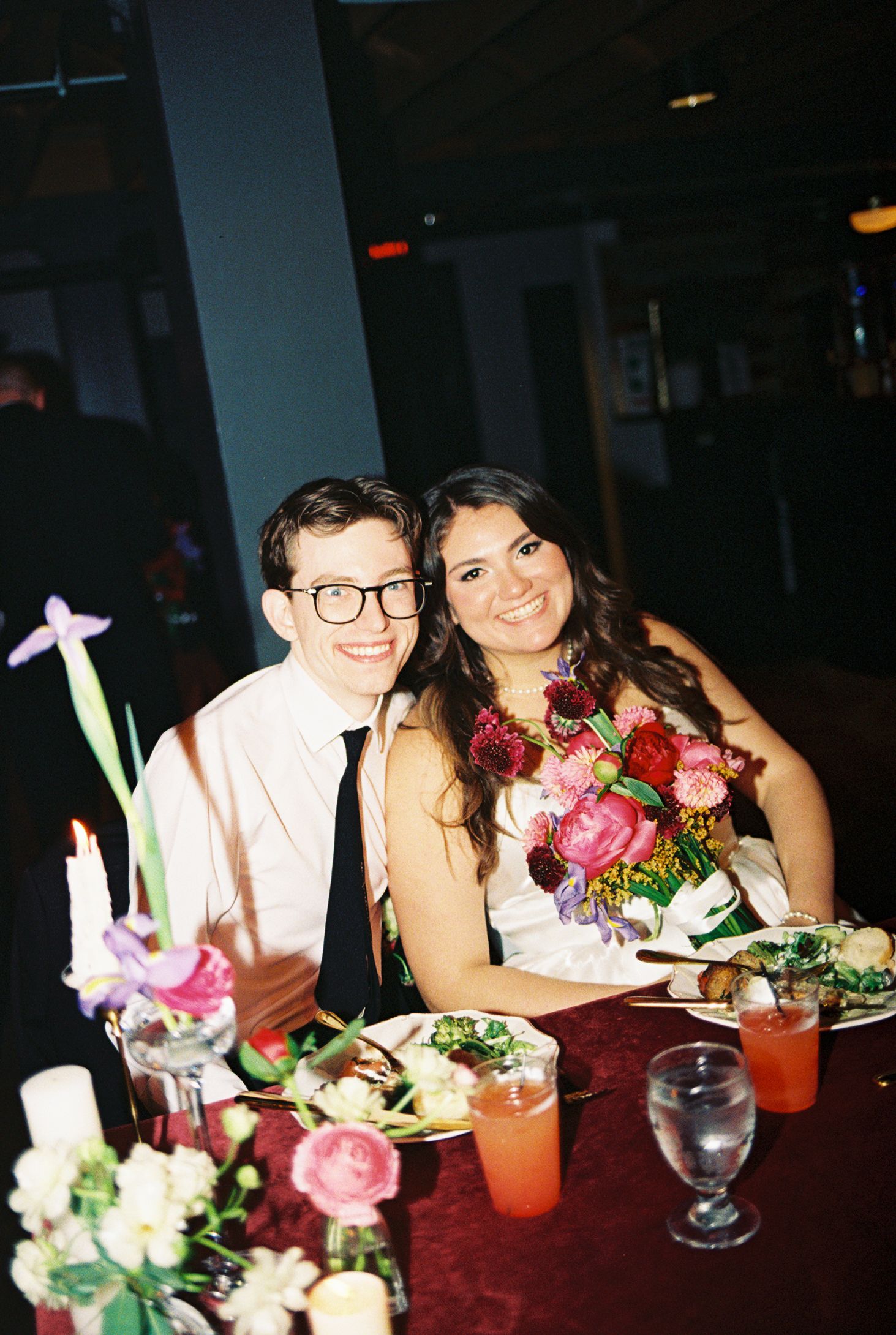 A smiling couple sits together at a floral-decorated wedding reception table with colorful bouquets and drinks.