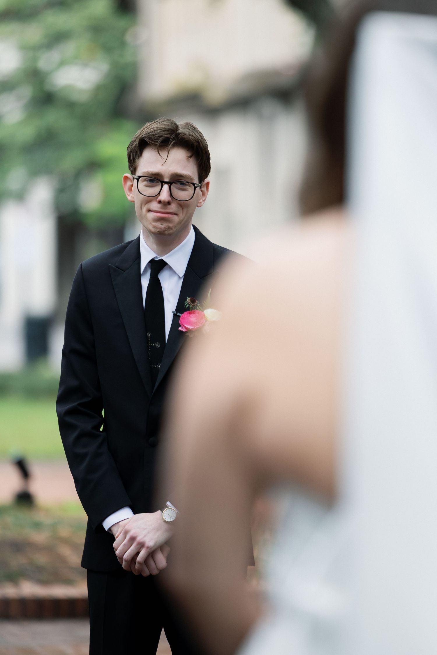Emotional groom in black suit with pink boutonniere watches bride approach during outdoor wedding ceremony.