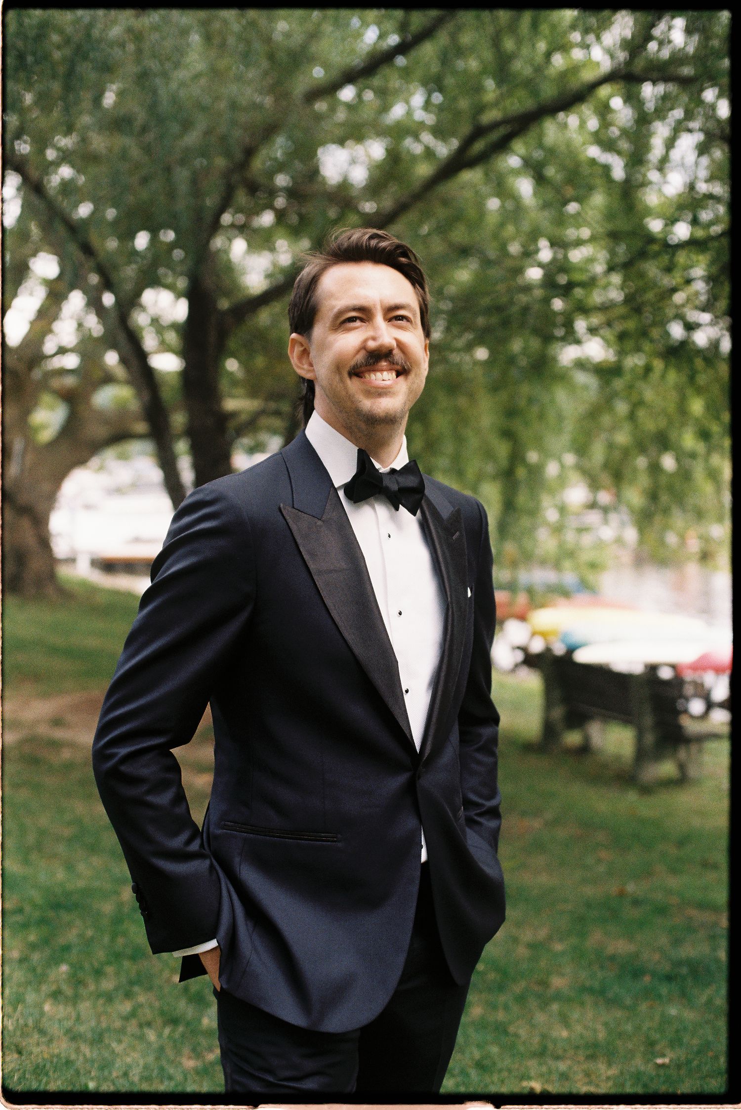 Handsome groom in navy tuxedo with bow tie smiles outdoors among green trees.
