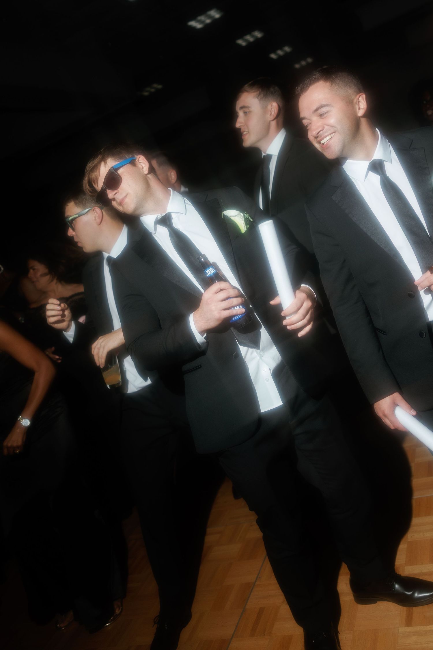 Groomsmen in black tuxedos laughing and celebrating together on the dance floor at a wedding reception.
