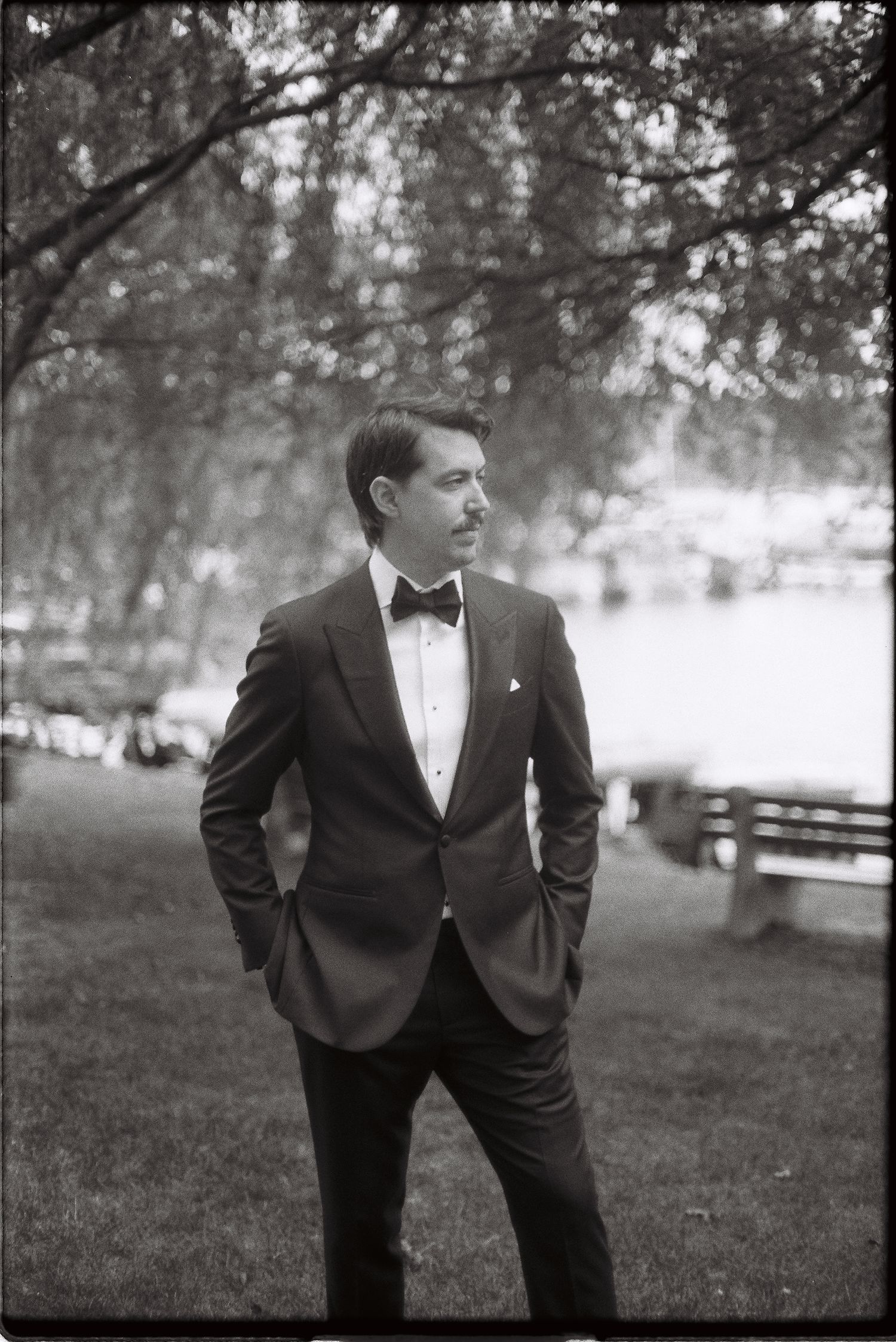 Elegant man in tuxedo with bow tie stands outdoors in park, black and white formal portrait photography.