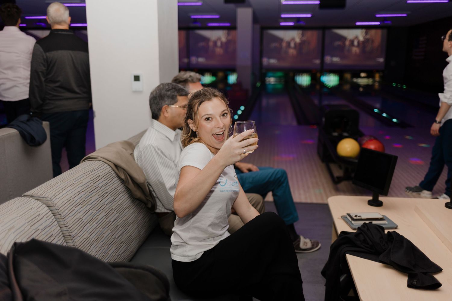 Several players follow through on their bowling form at a bowling alley illuminated by purple lighting.