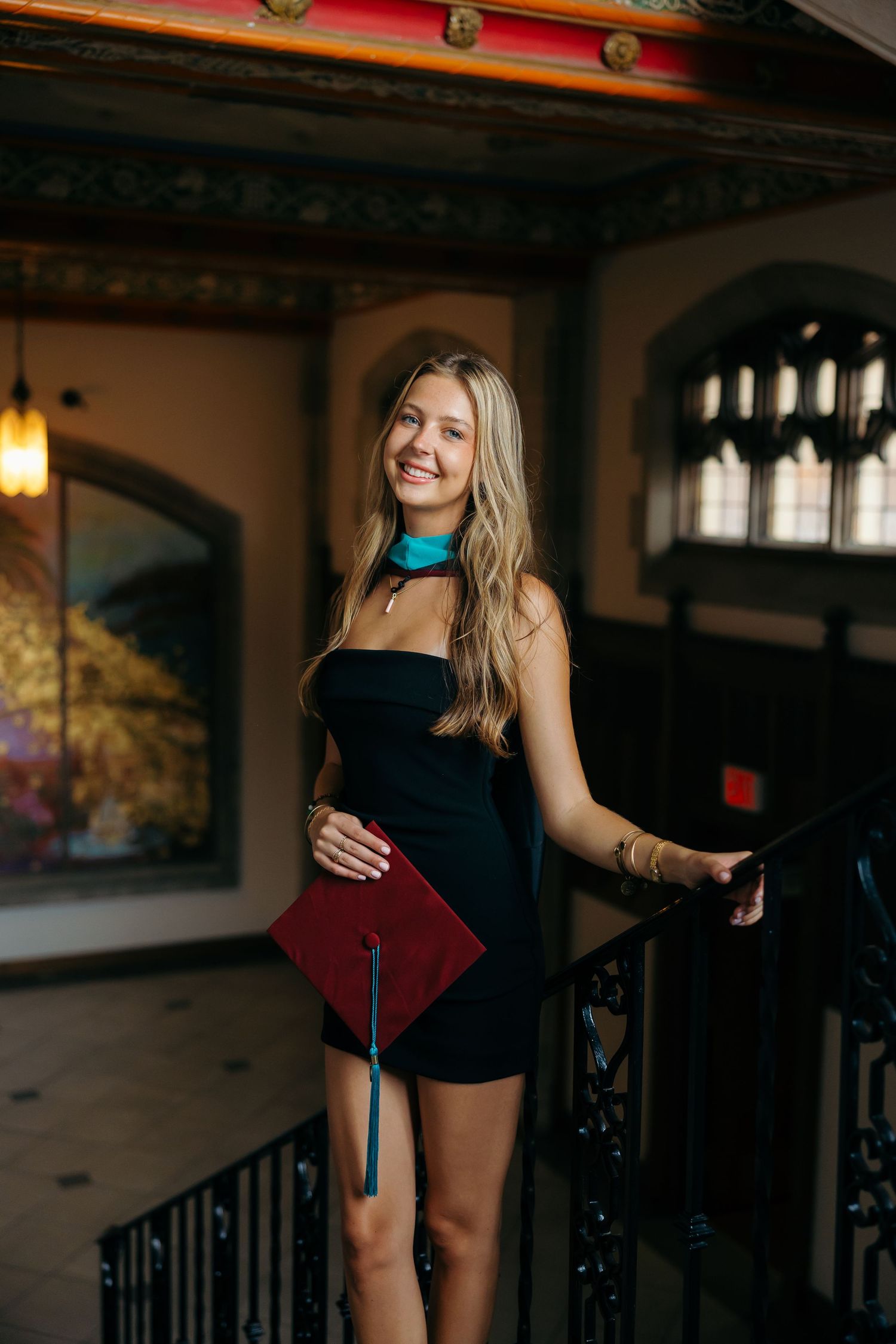 A graduate in a black dress poses with their diploma in an elegant indoor setting with ornate architecture.