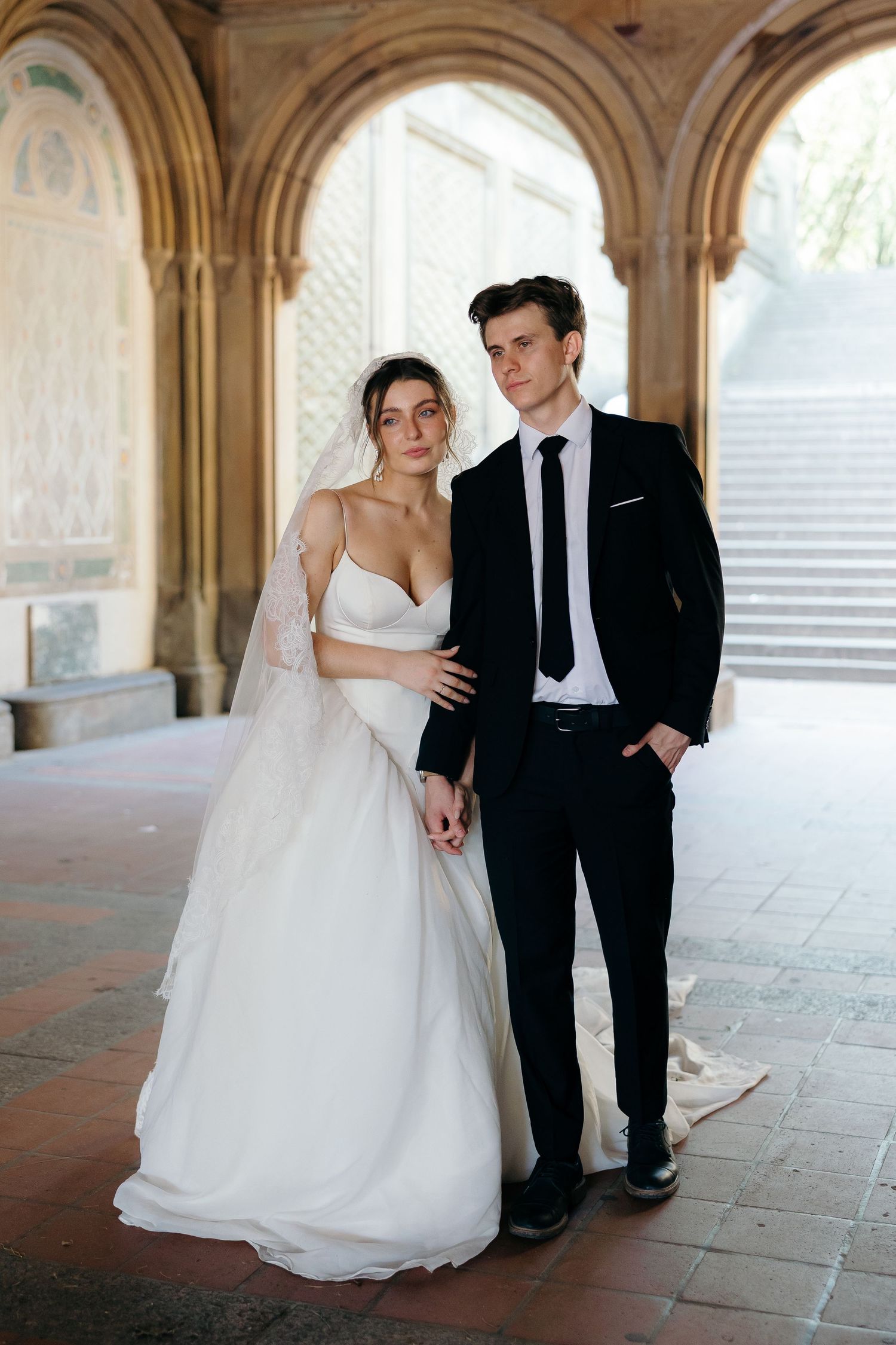 A bride in a flowing white dress and groom in black suit pose together under elegant stone arches.
