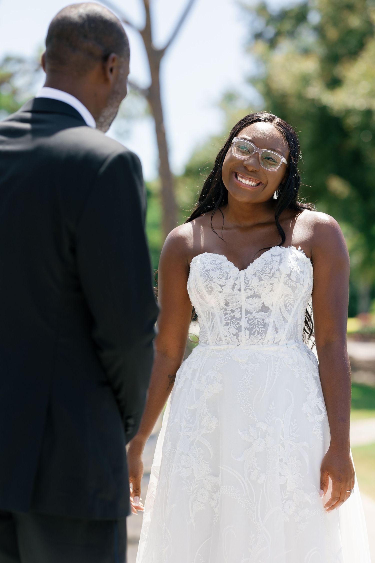 Smiling Black bride in strapless lace wedding gown and glasses faces groom in black suit outdoors.
