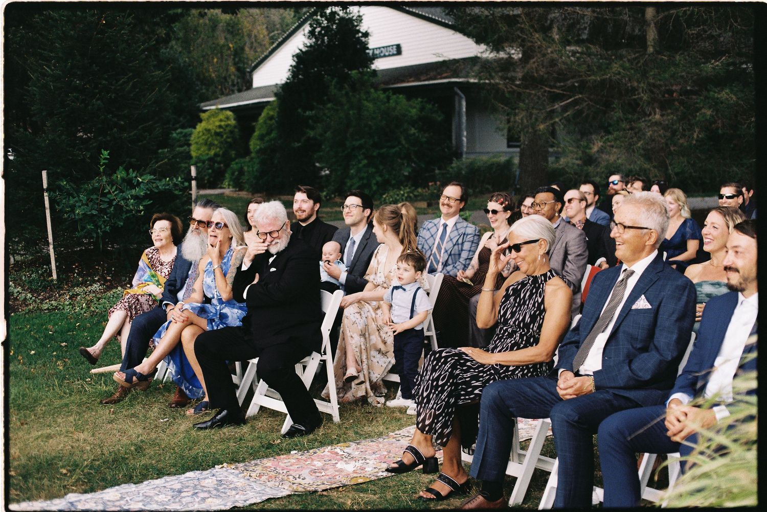 Guests seated in white chairs at an outdoor wedding ceremony on a sunny day surrounded by lush greenery.