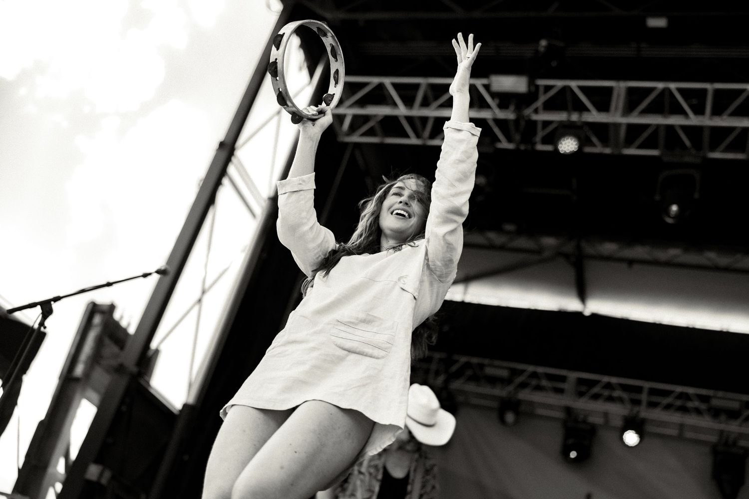 Black and white photo of a woman performing on stage, arms raised, holding a tambourine at an outdoor music festival.