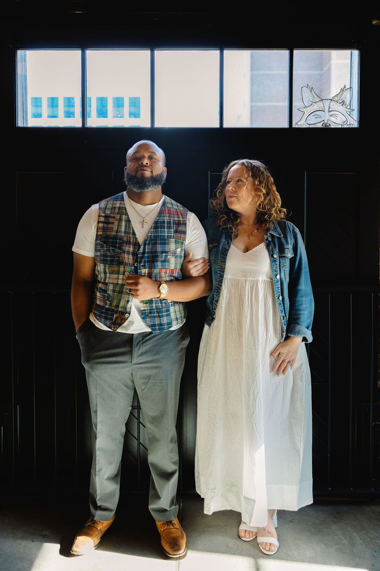 A couple stands together against a dark wall, with sunlight streaming through windows above them as they pose casually.