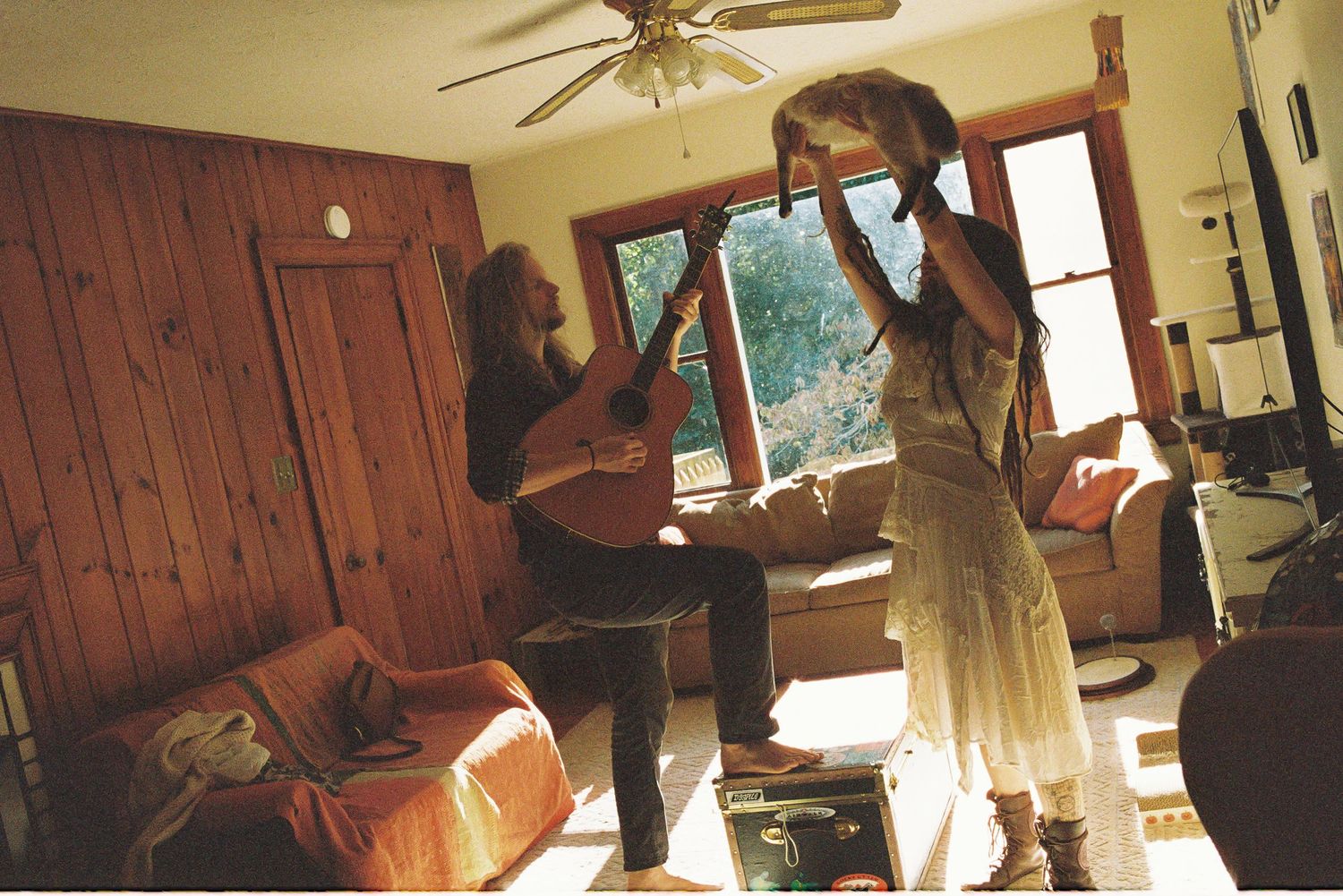 Two people in a sunlit wood-paneled living room, one playing acoustic guitar while the other dances in a white dress holding up a hat.