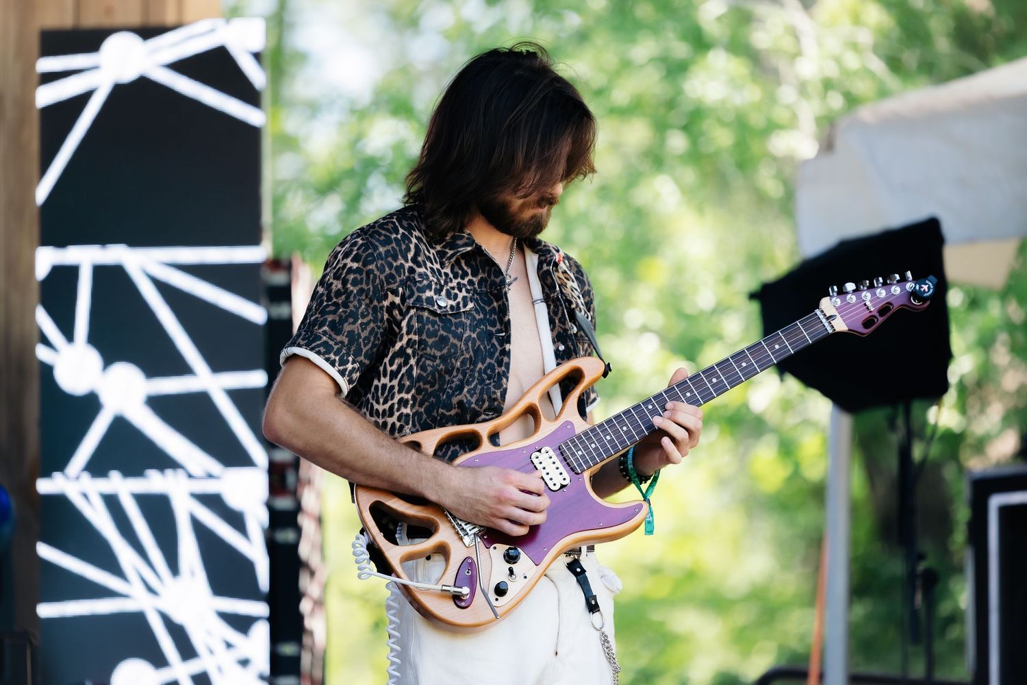 Guitarist performs on stage with pink electric guitar against geometric black and white backdrop during outdoor concert.