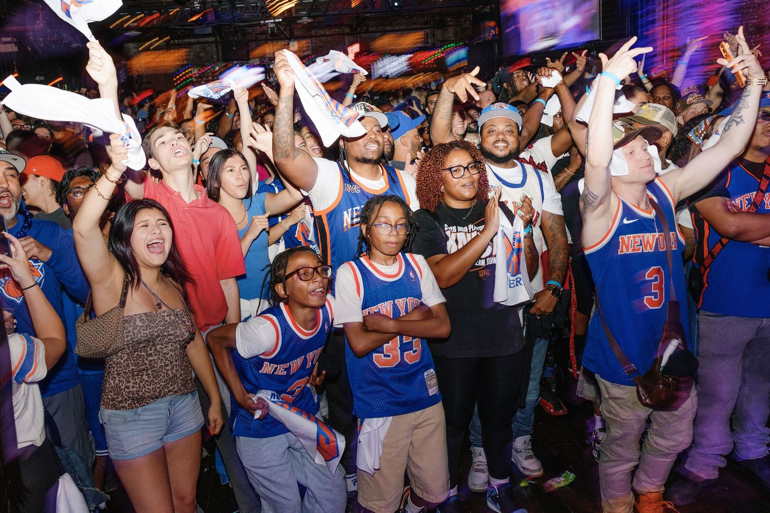 Excited New York Knicks fans in jerseys cheer at a crowded NBA viewing party event.