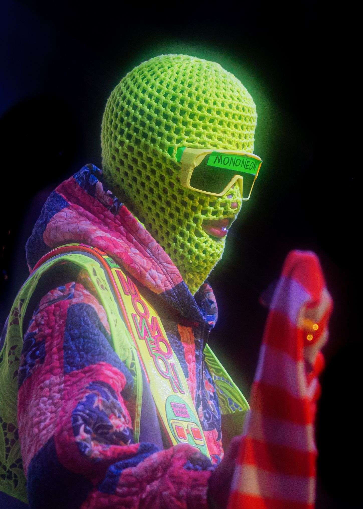 Person wearing neon green crochet ski mask and Moncler sunglasses with colorful jacket under UV light.