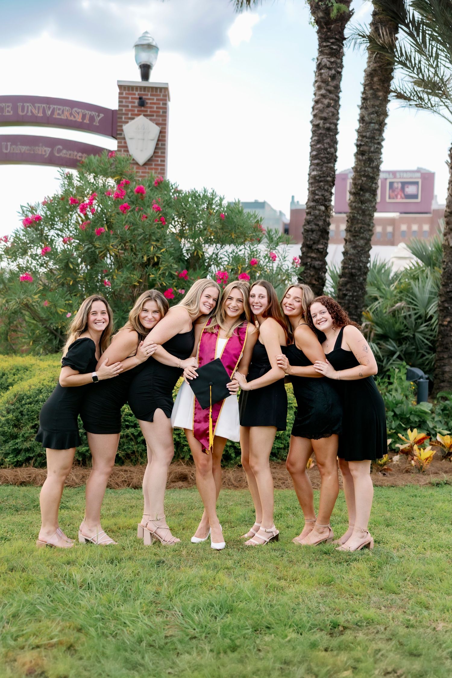 A group in black outfits poses together outdoors on grass near palm trees and flowering bushes at a university campus.