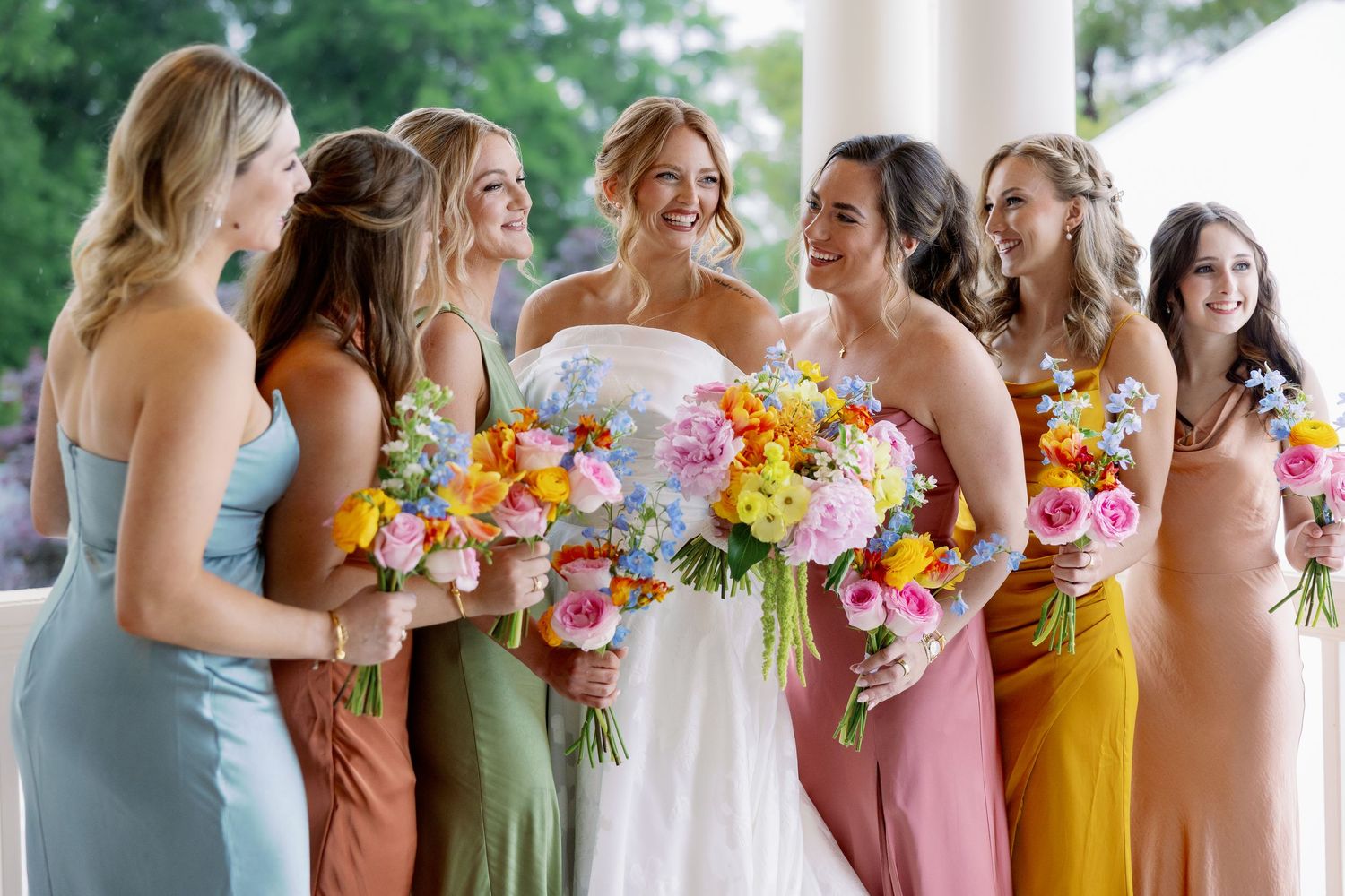 Bridesmaids in pastel and jewel-toned dresses stand together holding colorful floral bouquets at an outdoor wedding.