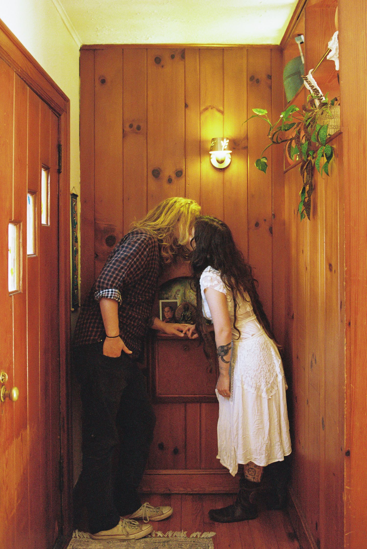 Two people with long hair kiss in a narrow wood-paneled hallway, one in a plaid shirt and sneakers, the other in a white dress and boots.