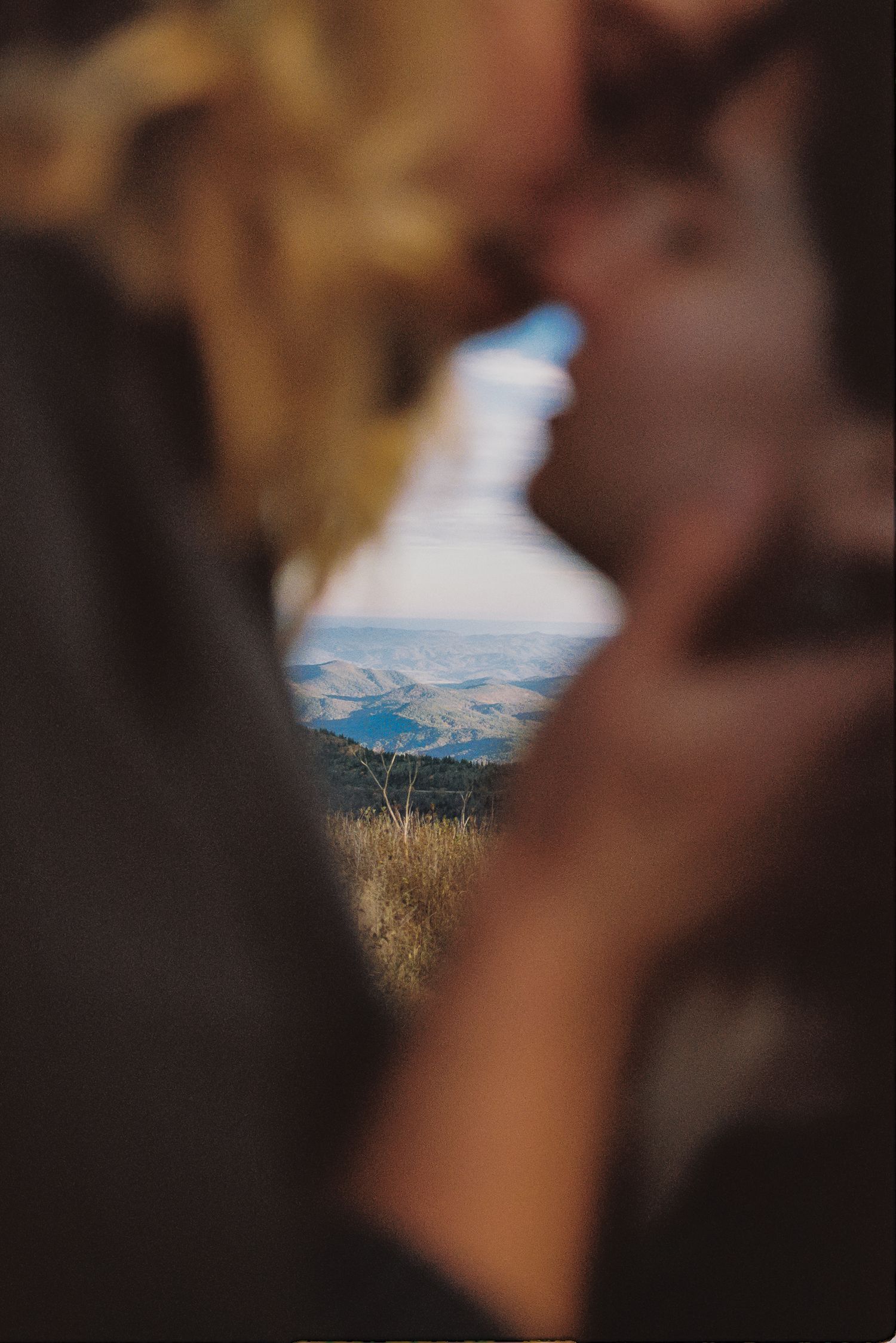 Couple sharing a romantic kiss in soft focus, framing a scenic mountain valley landscape in the background.