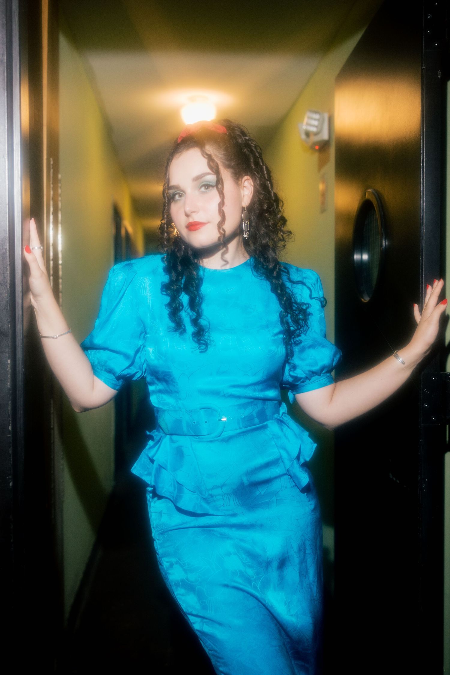 Woman in turquoise blue belted dress with braided hair poses in a dimly lit hallway doorway.