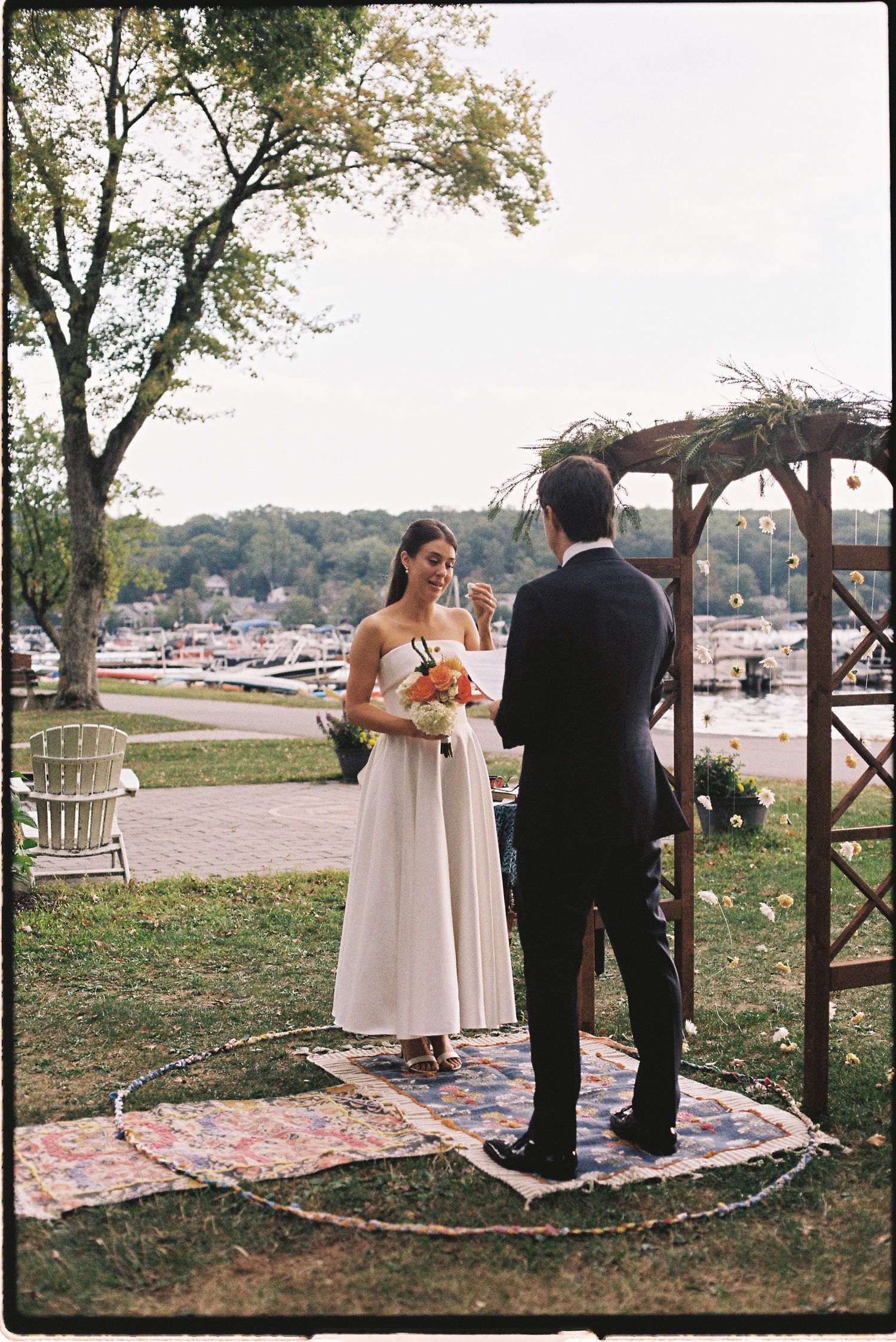 Bride and groom exchange vows outdoors near a lakeside arbor, bride holding a colorful bouquet in a white gown.