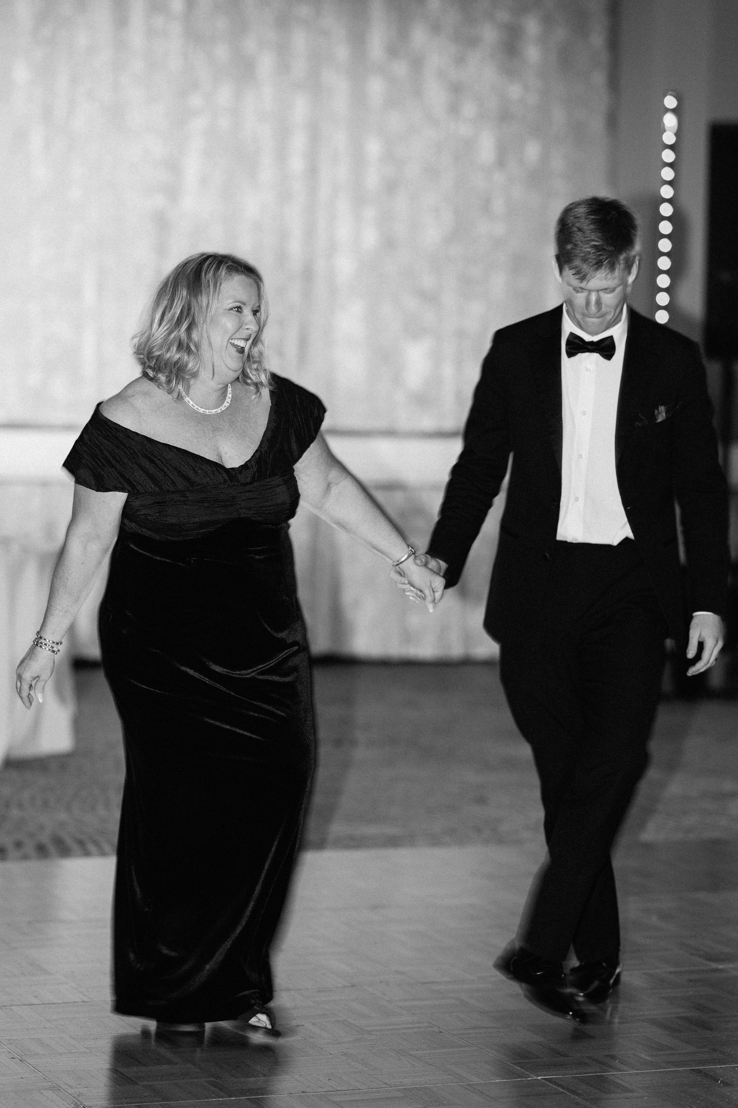 Elegant couple in formal black attire holding hands and smiling while walking on a dance floor at a wedding reception.