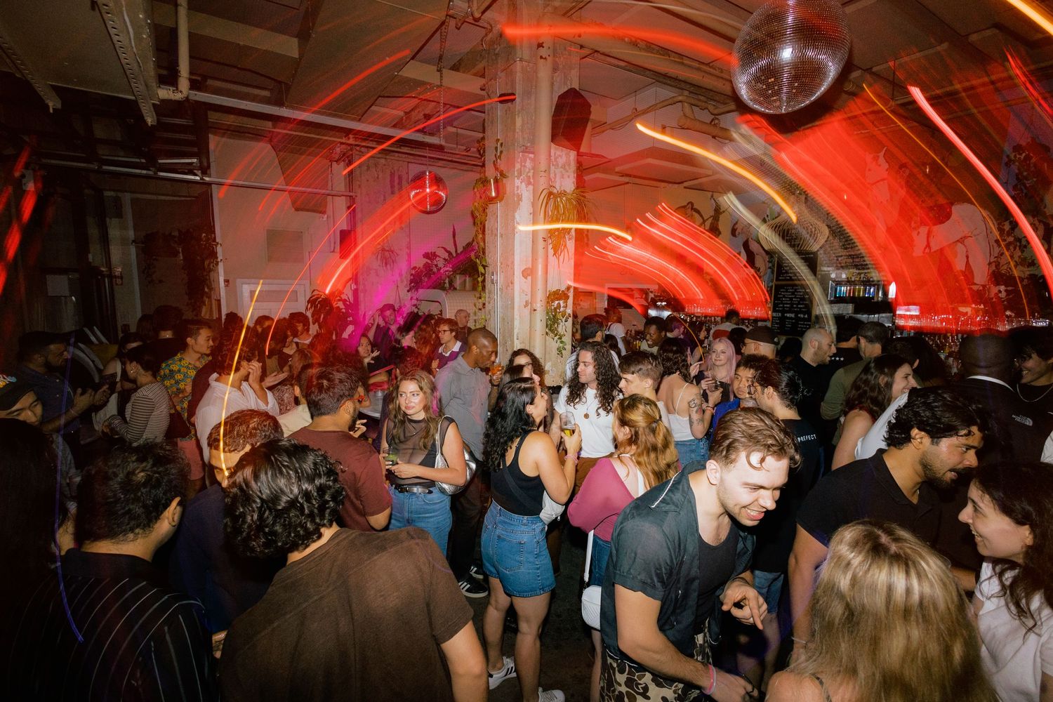 Crowded nightclub dance floor with red lighting and disco ball, people dancing and socializing at a lively party event.