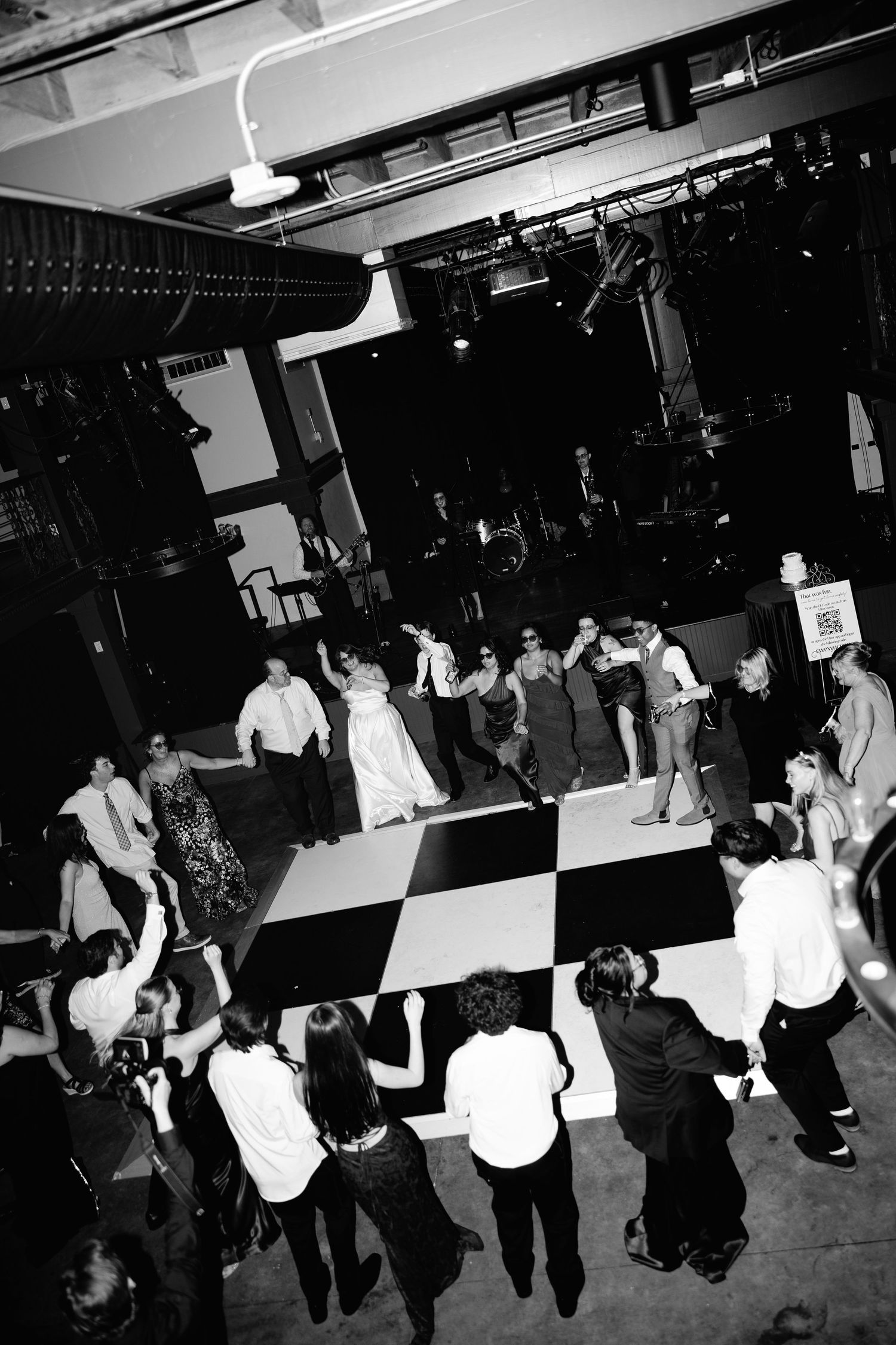 Black and white aerial view of wedding guests dancing on a checkered dance floor at a reception venue.