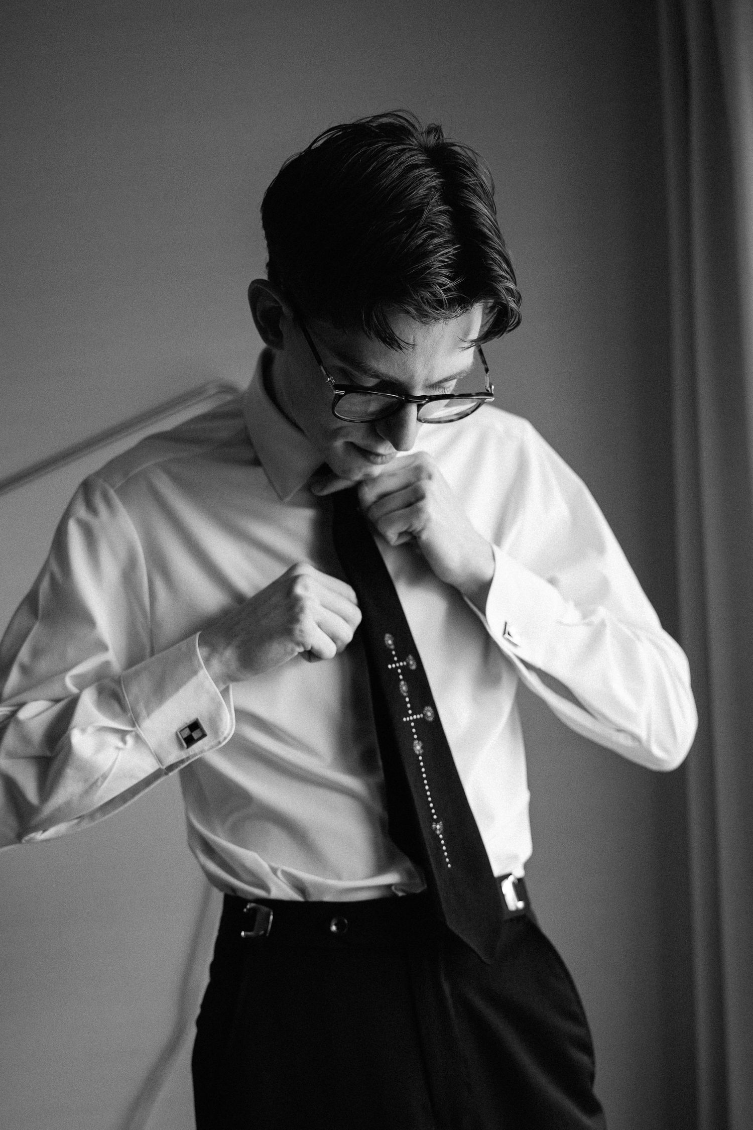 Black and white photo of a young man in white dress shirt adjusting his tie, wearing glasses and cufflinks.