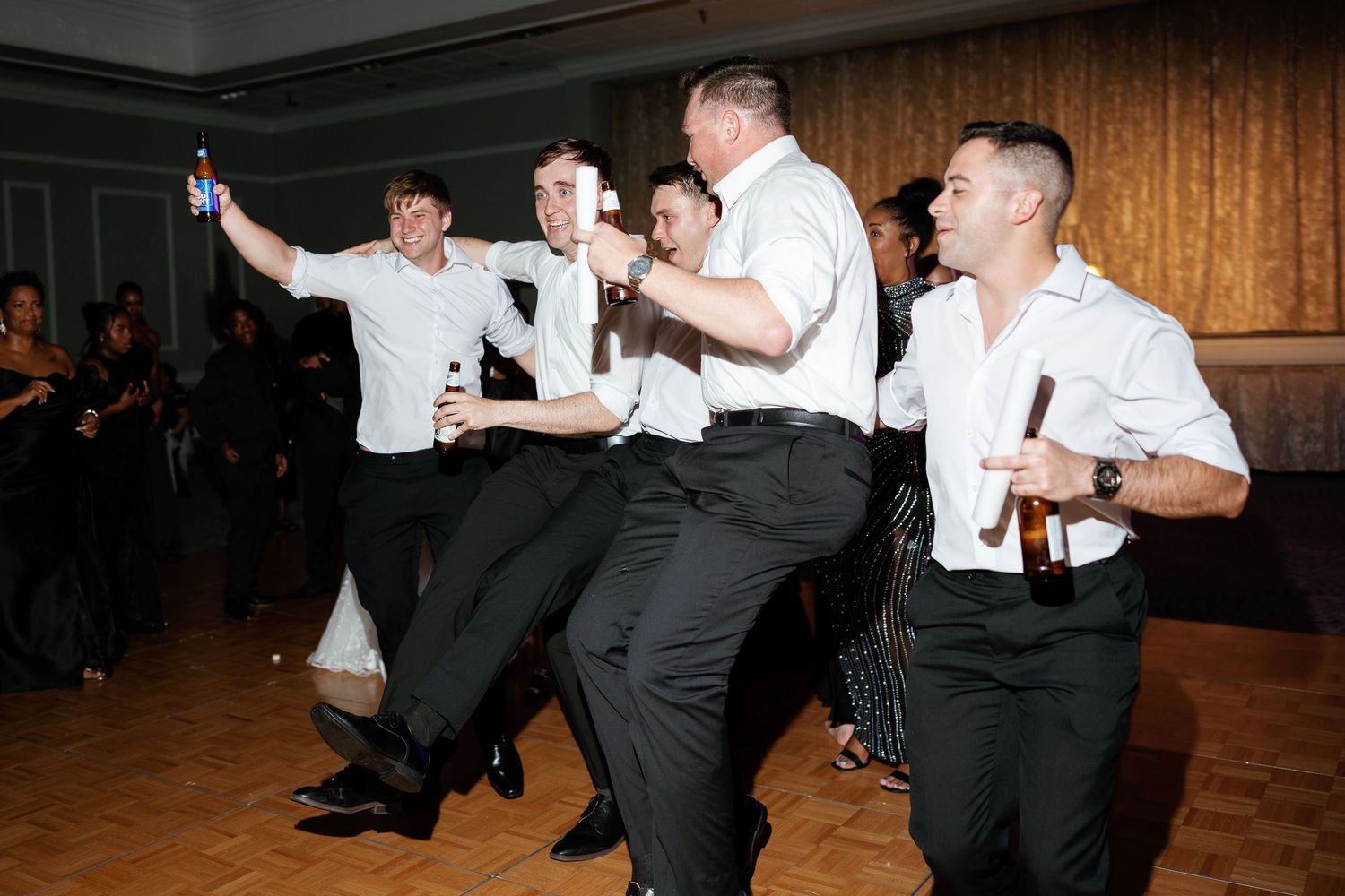 Men in white shirts and black pants dancing energetically at a formal event, holding beer bottles on a dance floor.
