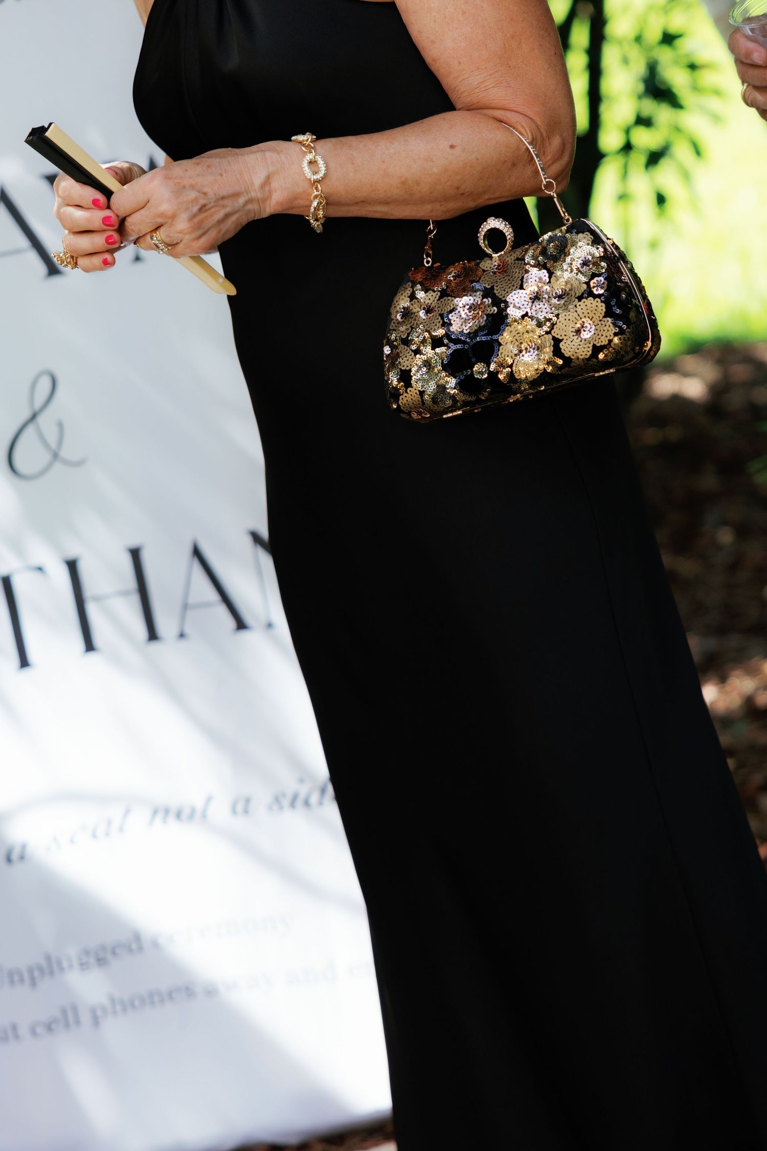 Woman in black gown holding decorative sequined clutch purse at outdoor wedding ceremony.