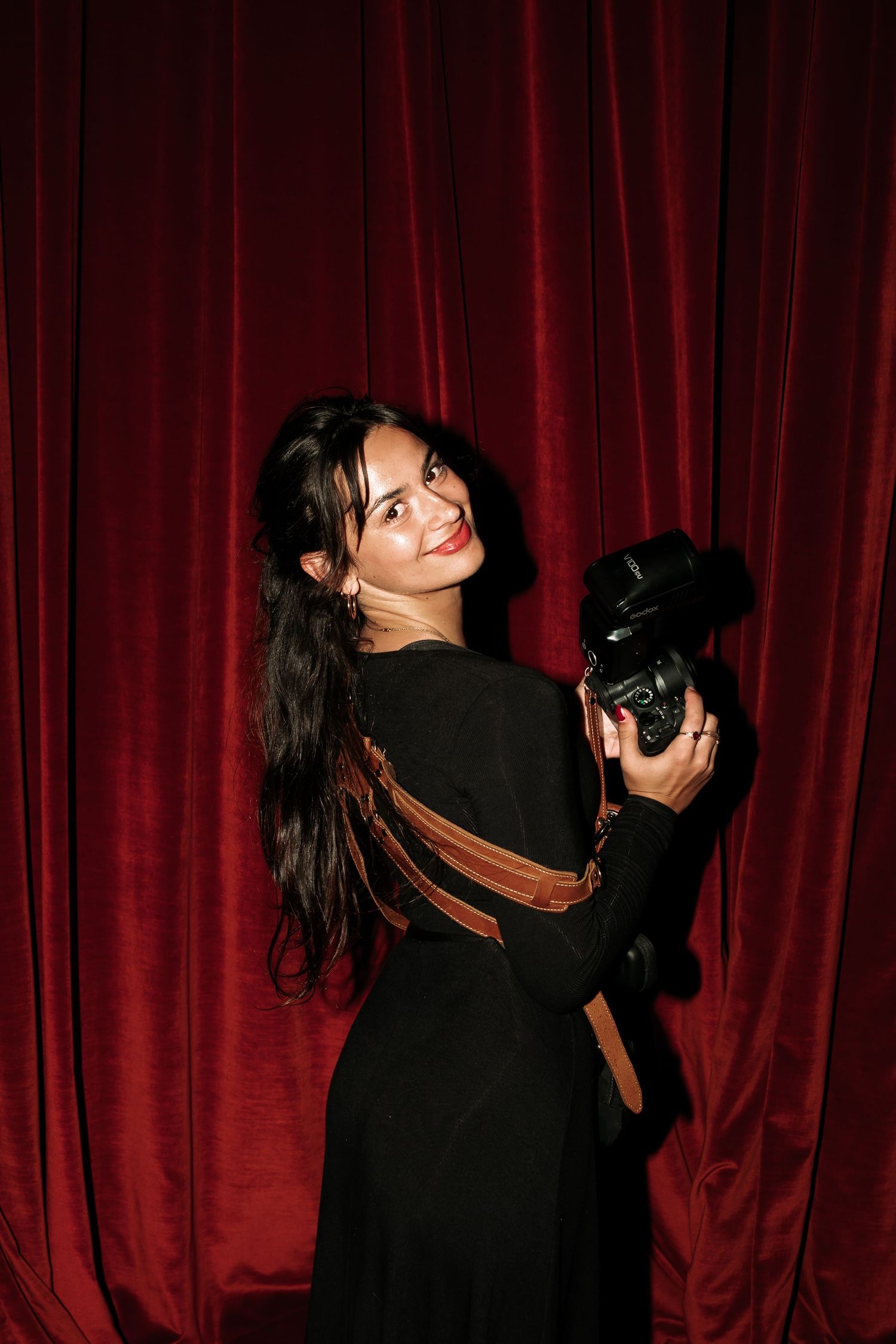 Smiling woman in black dress holding a camera, posing in front of red velvet curtains.