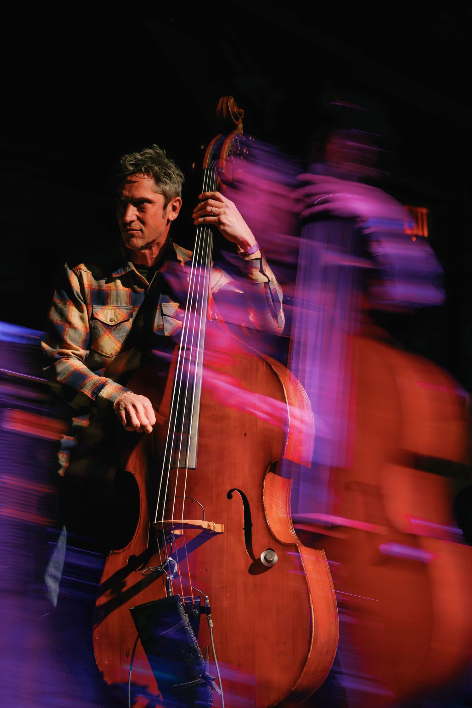 Motion blur captures a jazz bassist performing on upright bass with dramatic purple and red stage lighting.