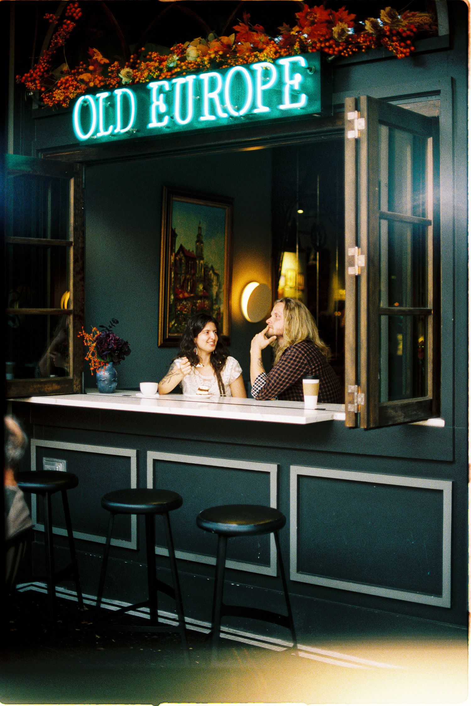 Two people chat over coffee at a cozy window counter under a glowing Old Europe neon sign, with bar stools and autumn floral decor.