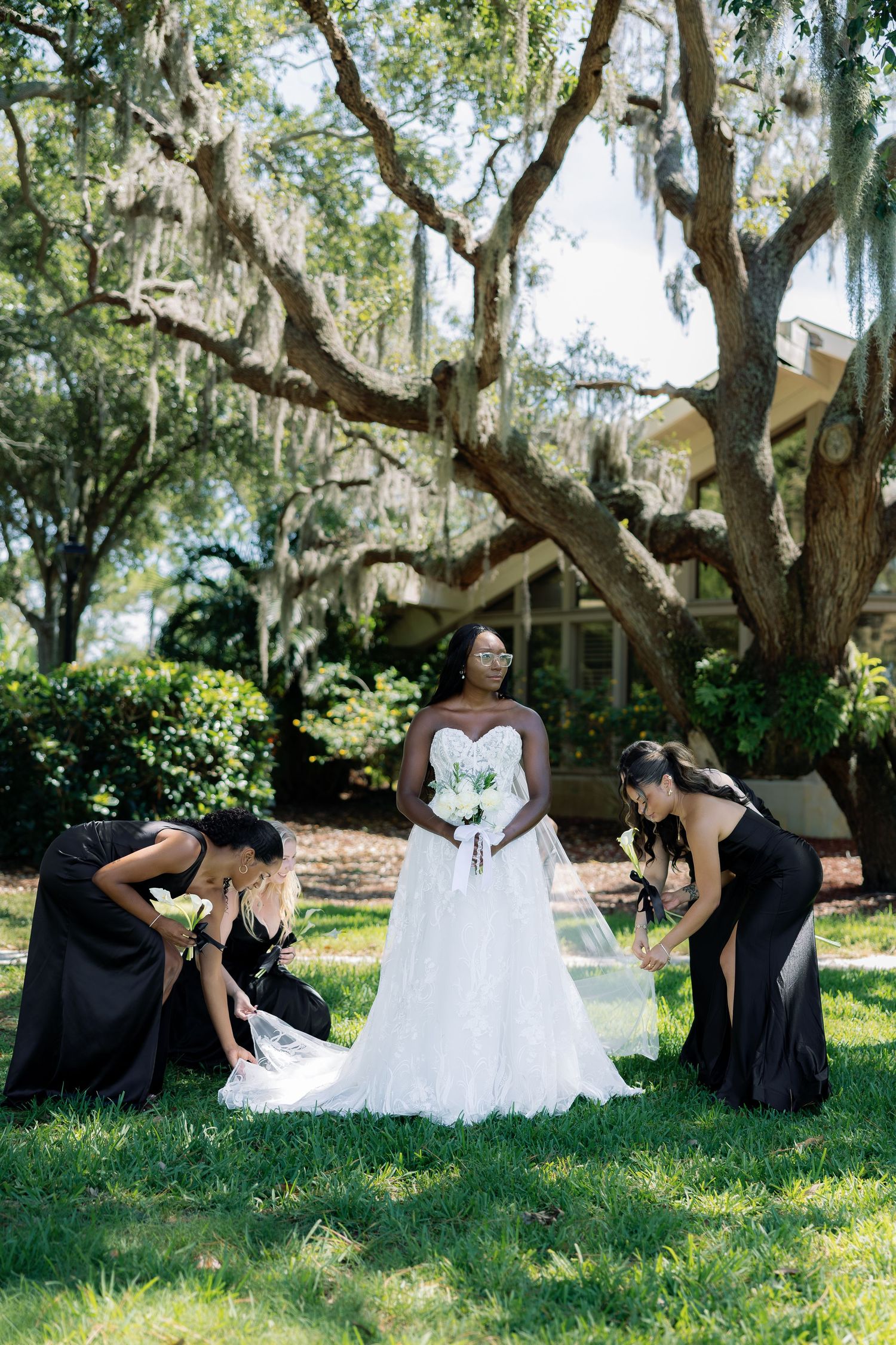 Bride in white strapless gown holds bouquet while bridesmaids in black dresses adjust her veil outdoors.
