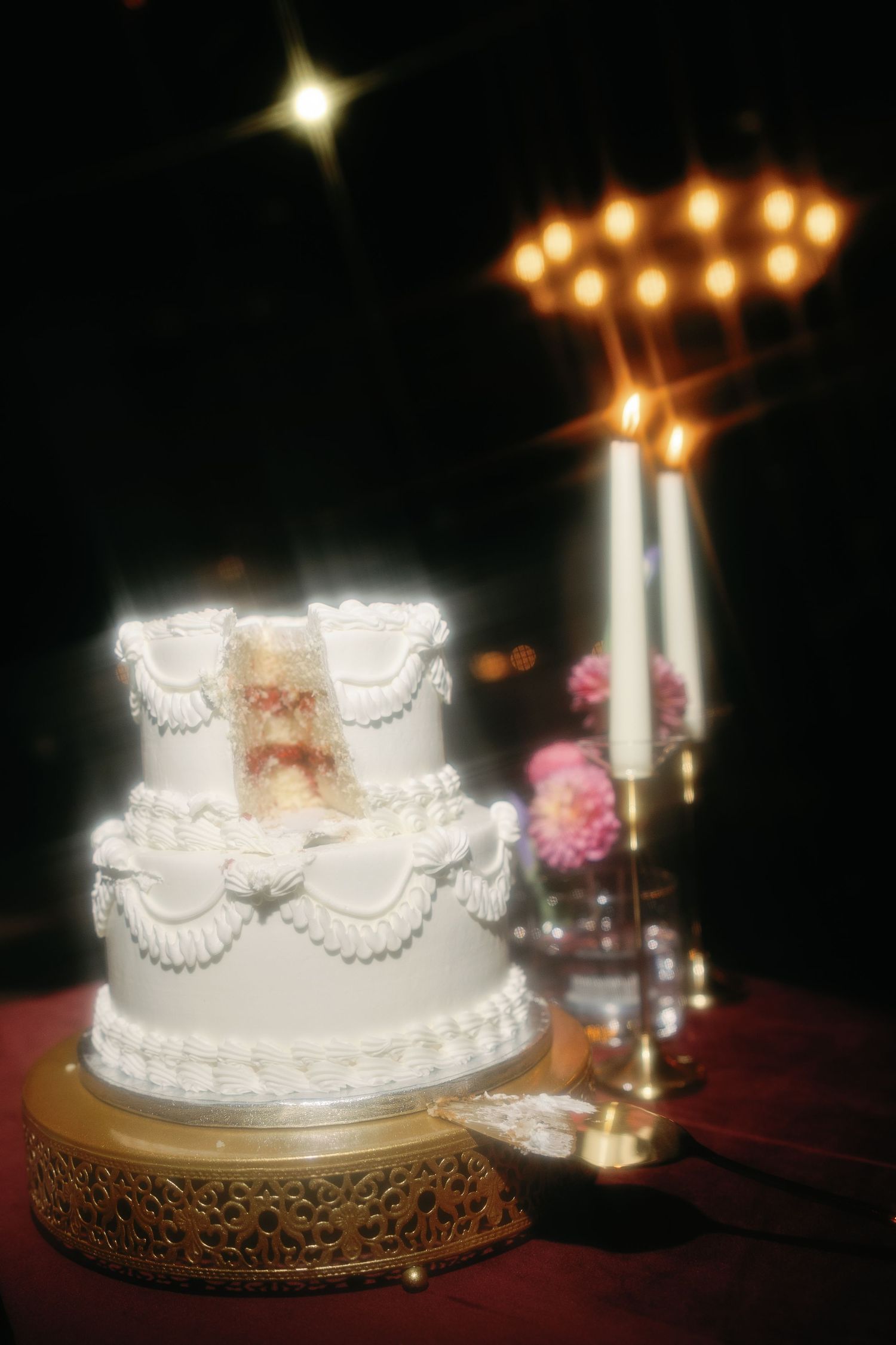Elegant two-tier white wedding cake with lace frosting on gold stand, beside lit candles and flowers in dark reception setting.