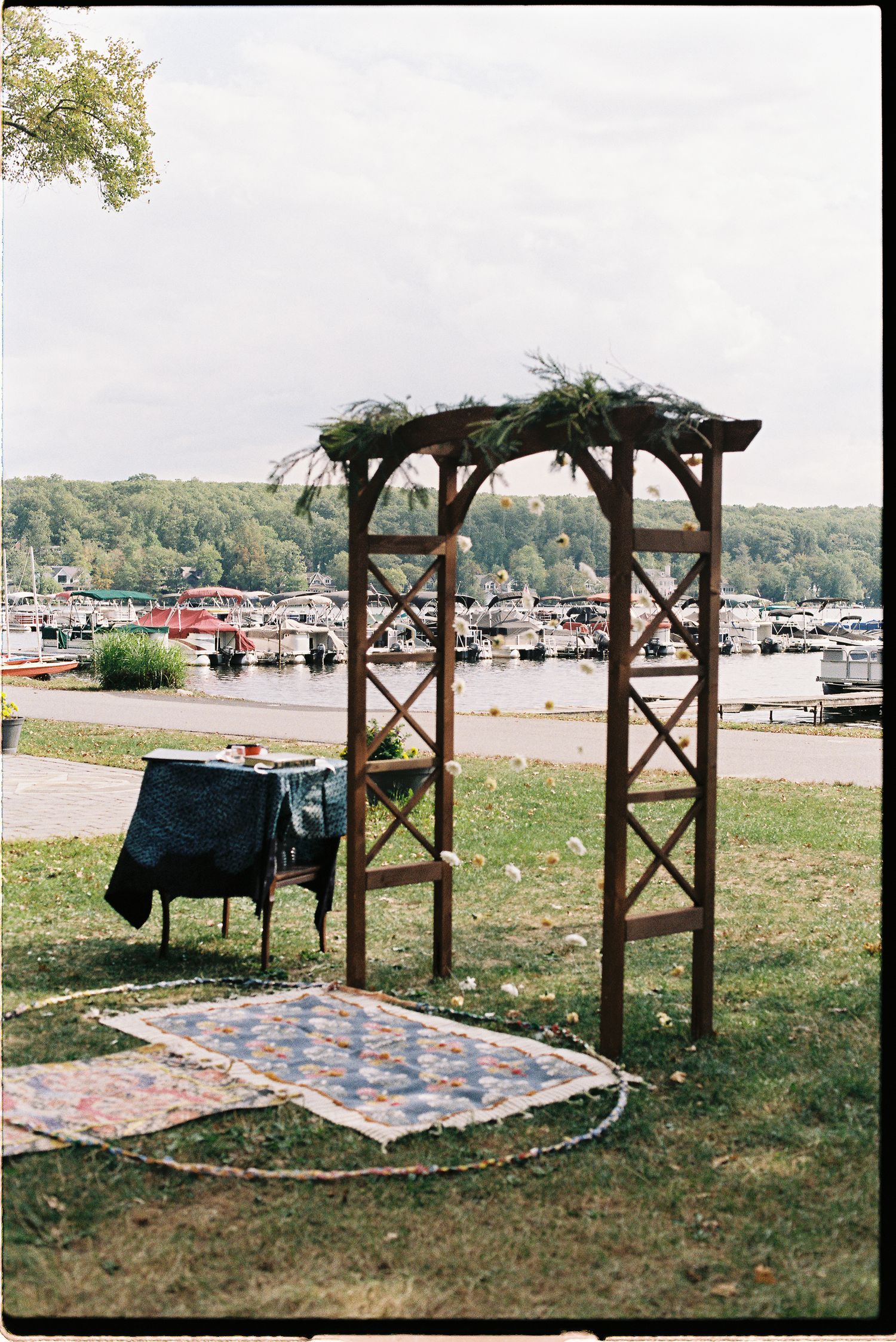 Wooden wedding arch decorated with greenery overlooks a marina, with colorful quilts laid on grass nearby.