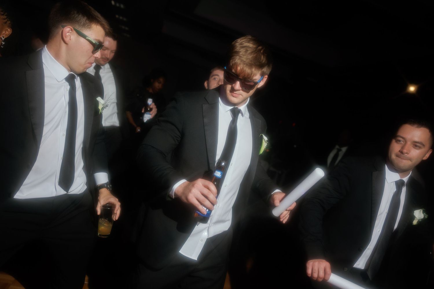 Groomsmen in black suits and sunglasses holding glow sticks celebrate at evening wedding reception.