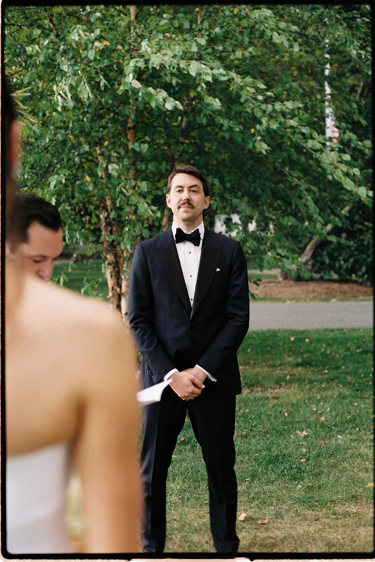 Groom in black tuxedo and bow tie watches bride approach during outdoor wedding ceremony on green lawn.